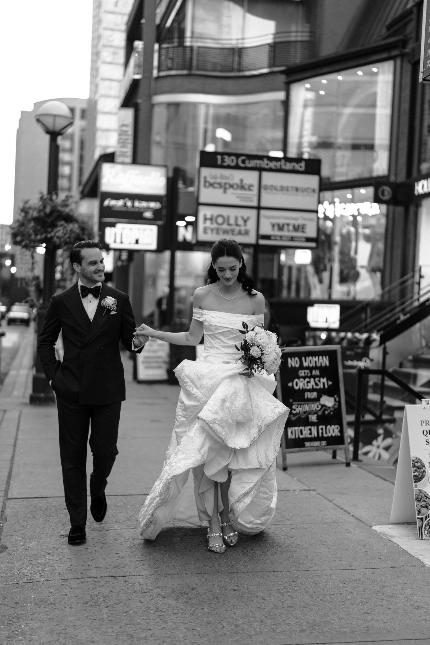 A couple in wedding attire walks down a city sidewalk with urban storefronts and signs visible in the background.