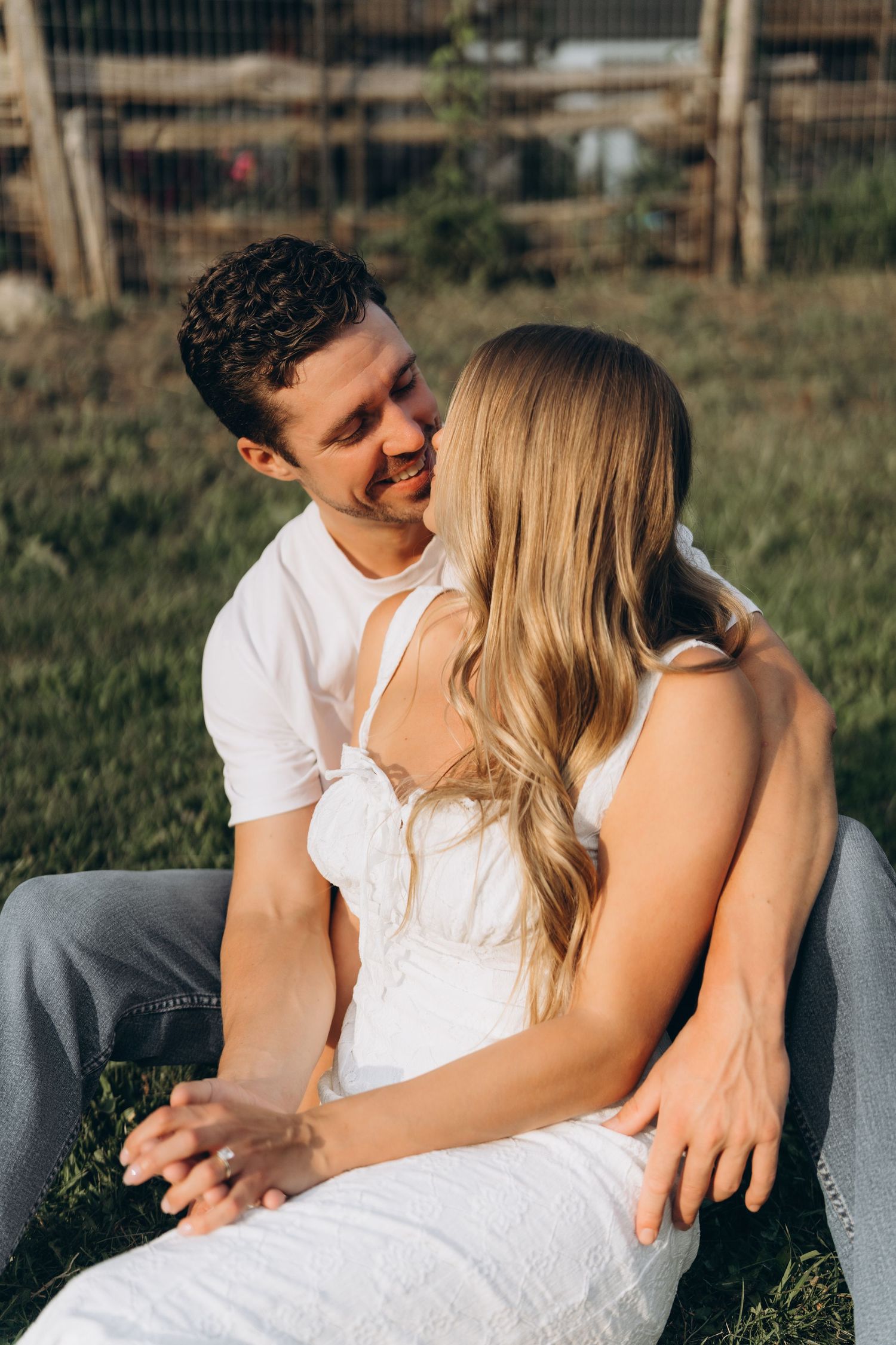 A couple in white outfits sharing an intimate moment while sitting outdoors in a grassy field.