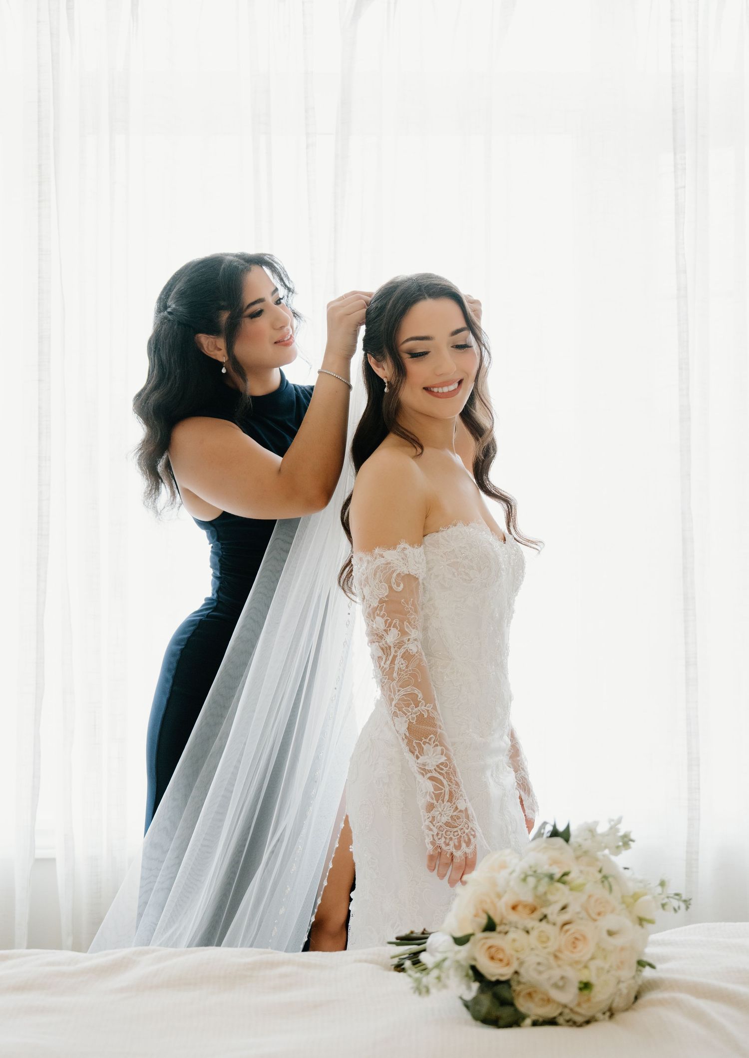 A bride getting ready in a white lace gown with long sleeves as her veil is being adjusted near bright windows.