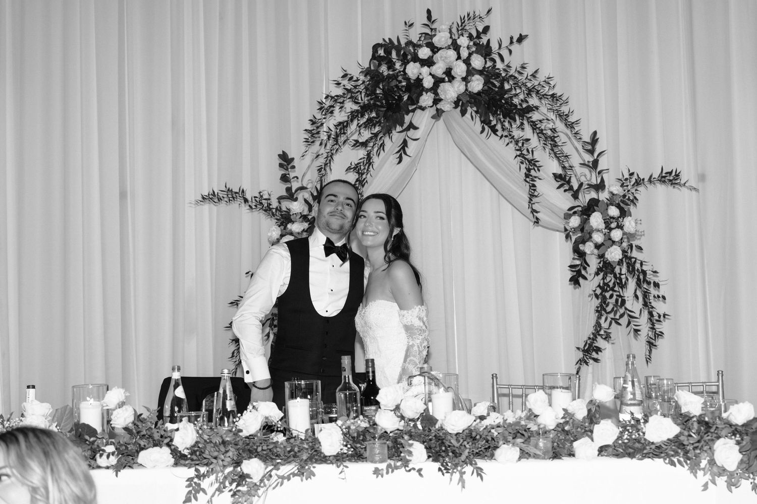 A black and white wedding reception photo showing tables with floral centerpieces and guests under elegant draping.
