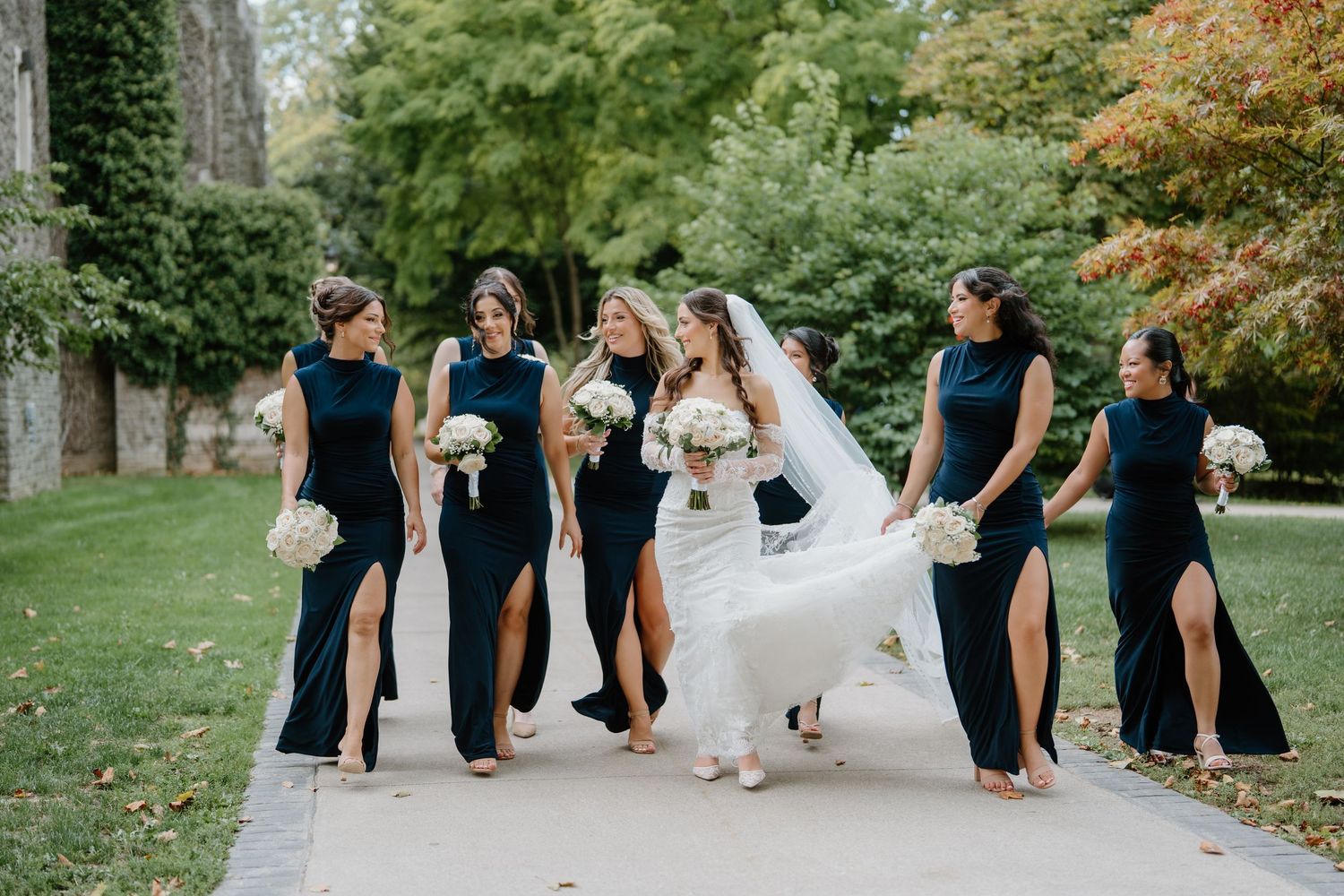 Wedding party walks in a line along a garden path wearing black and white formal attire during an outdoor celebration.