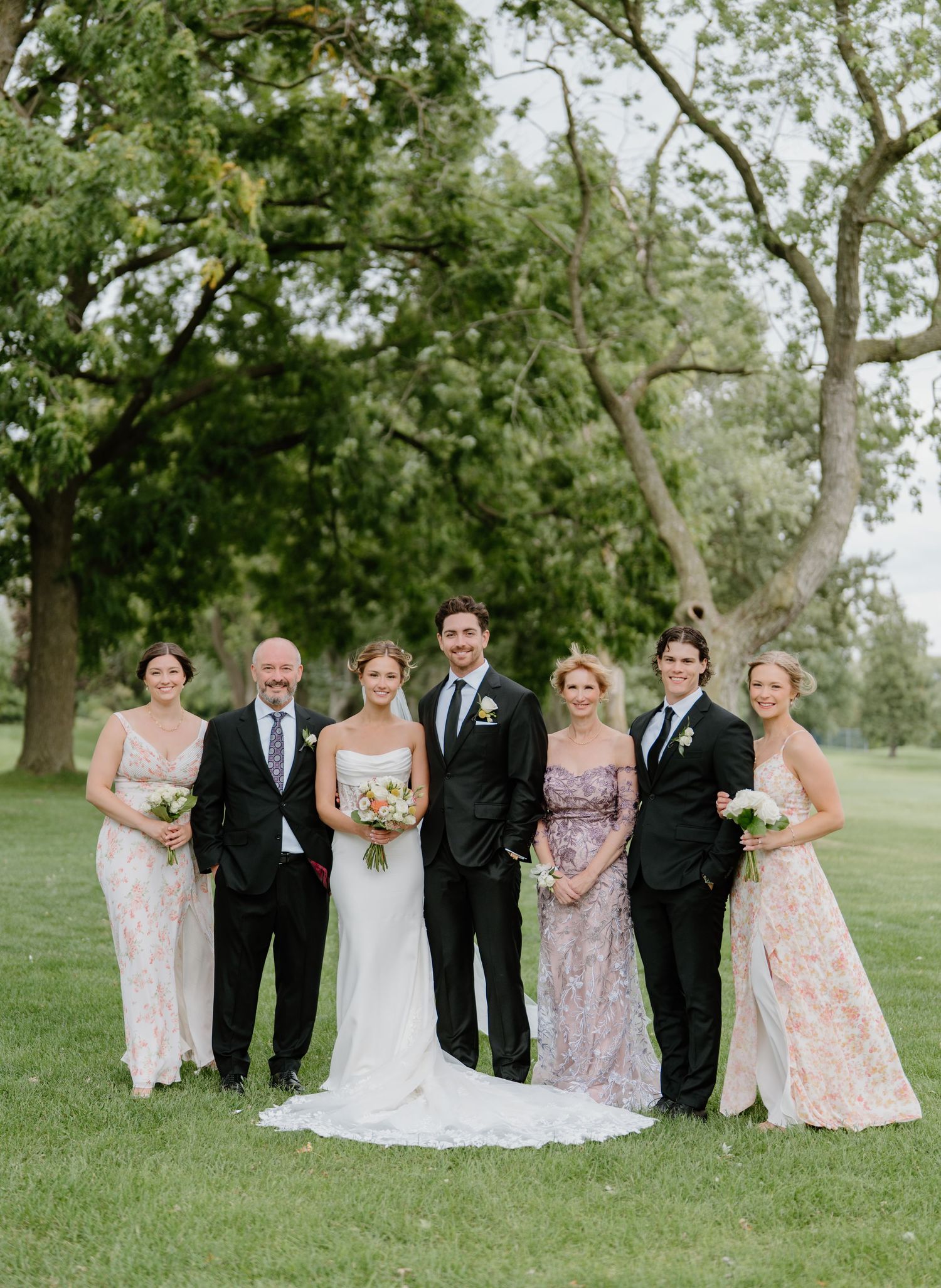 Wedding party poses together on a grassy lawn with trees in the background wearing formal attire in pastel colors.