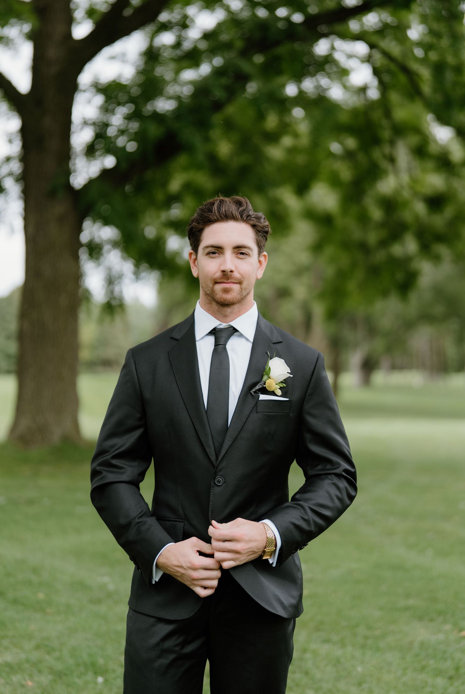 A groom in a classic black suit and tie adjusts his cuffs while standing in a park setting.