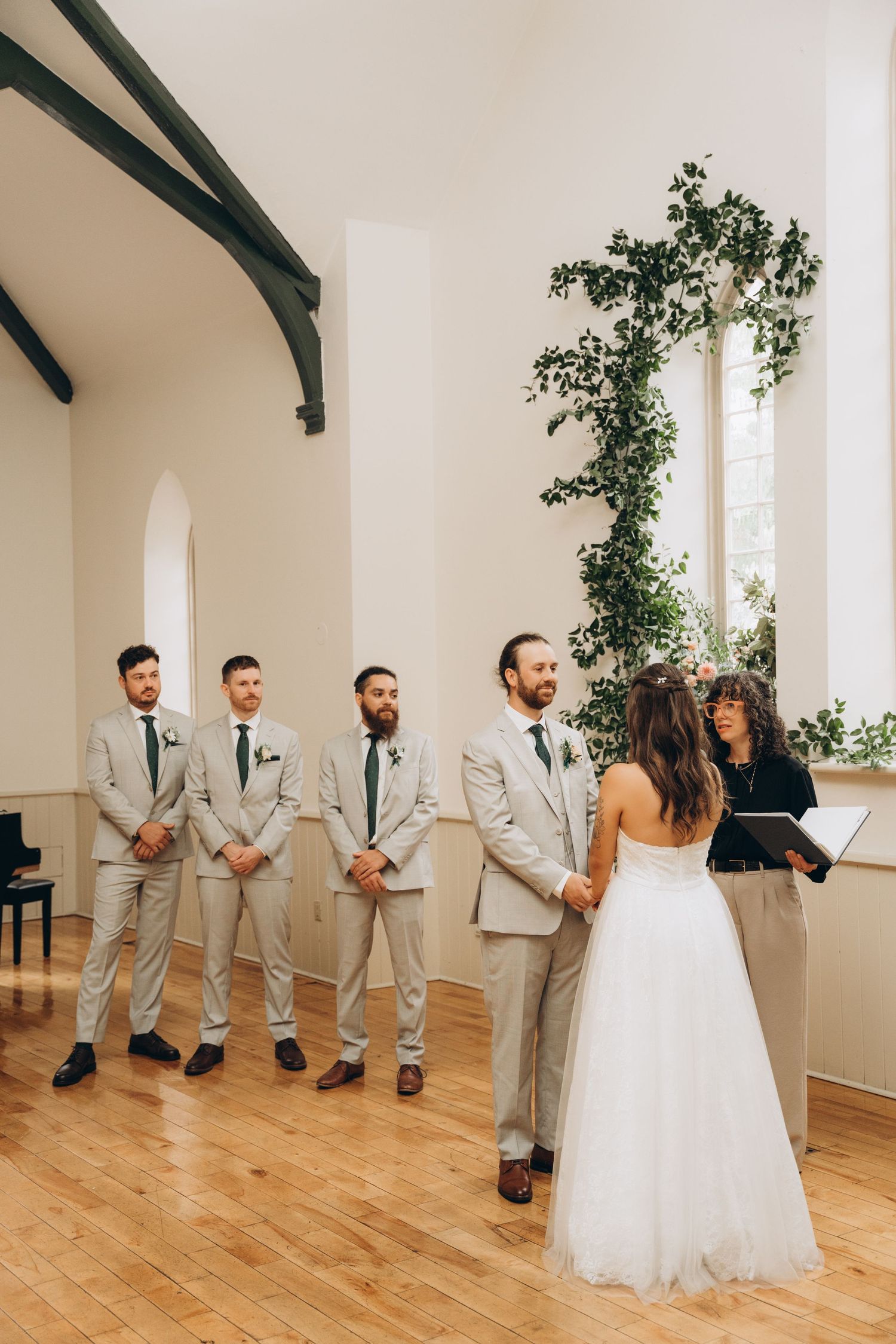 Wedding party in light gray suits stands against white wall while couple shares intimate moment in elegant venue.