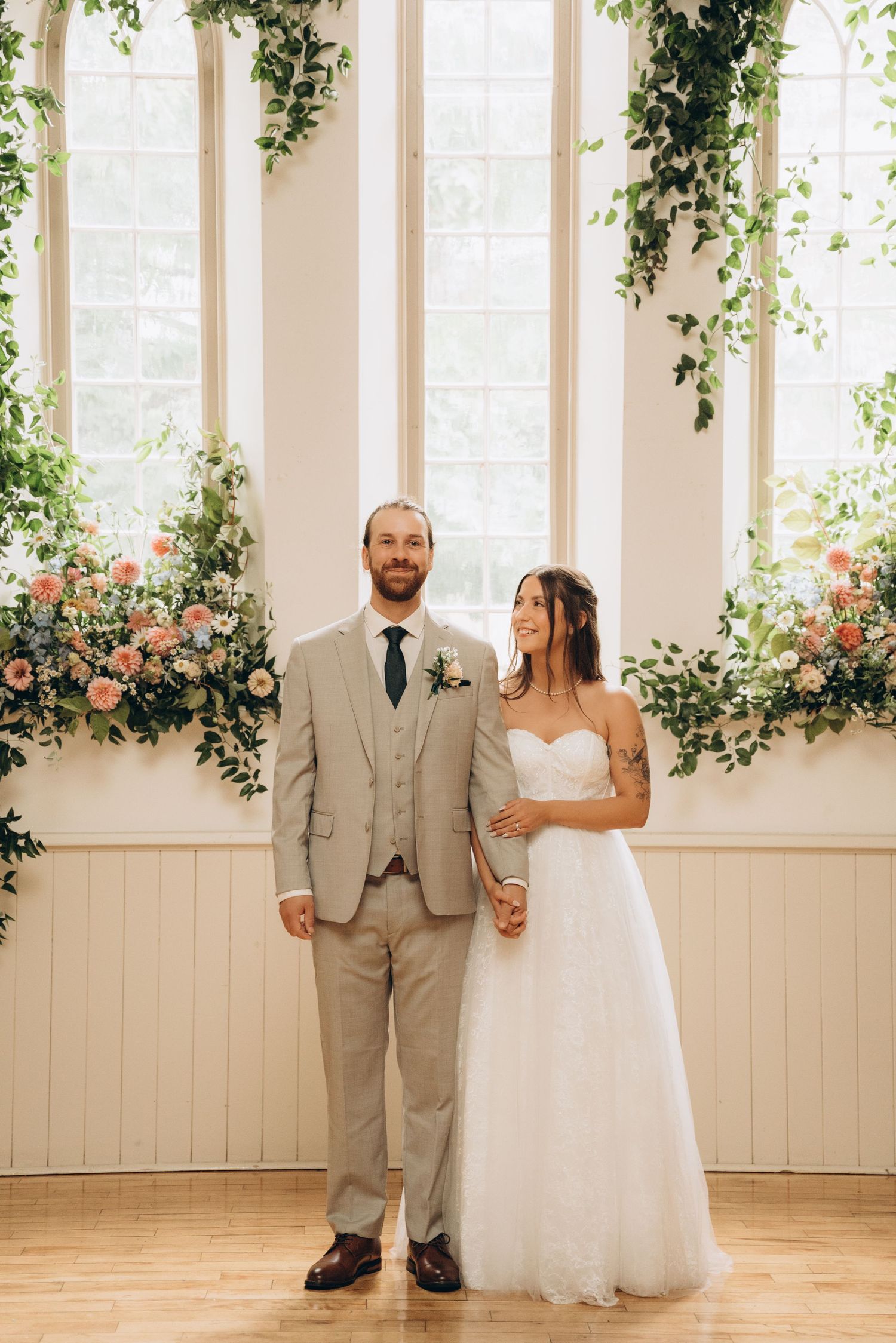 A bride and groom pose in a sunlit room decorated with climbing vines and pink roses against white walls.