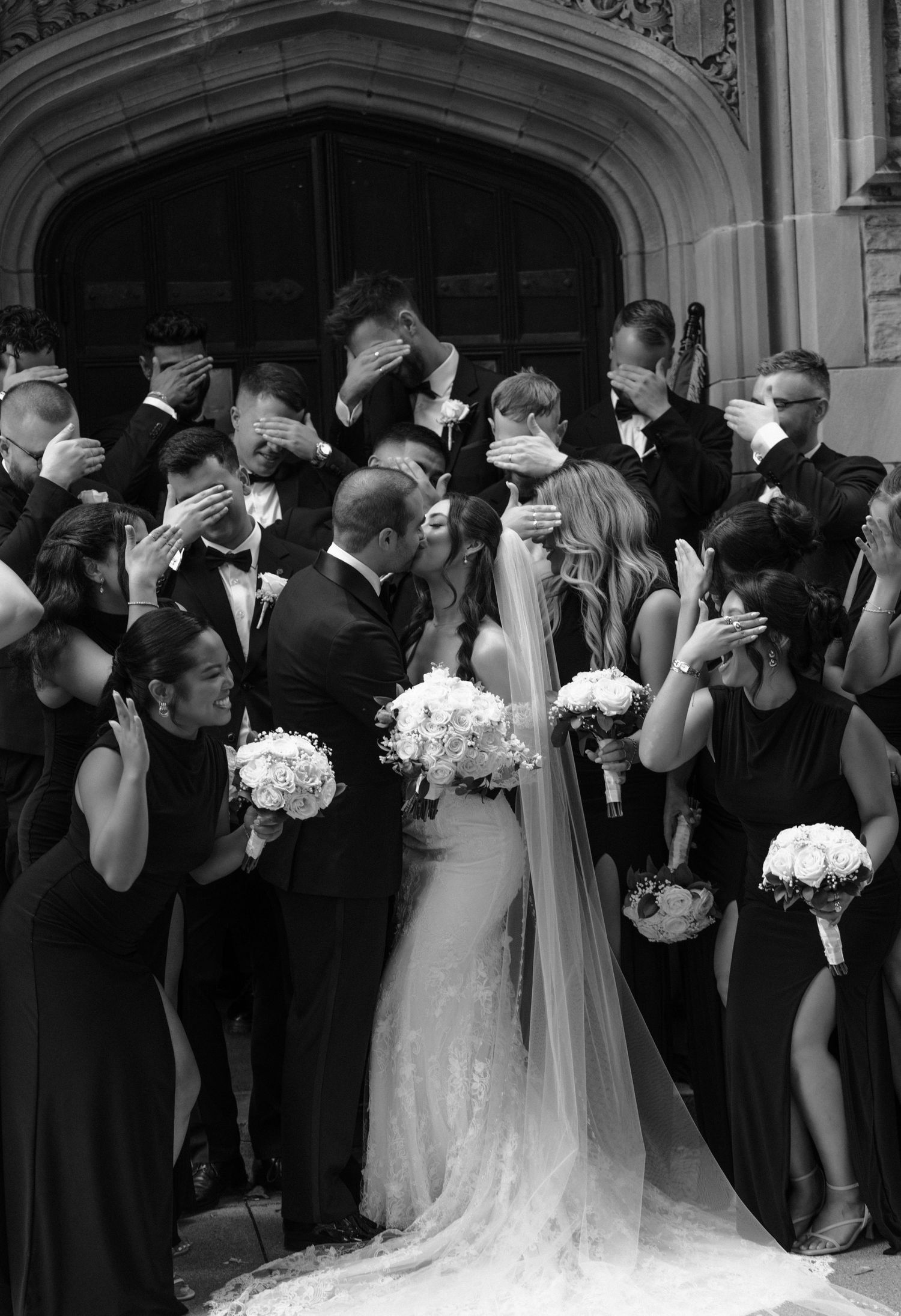Wedding party members in formal black and white attire share kisses in an elegant arched doorway.