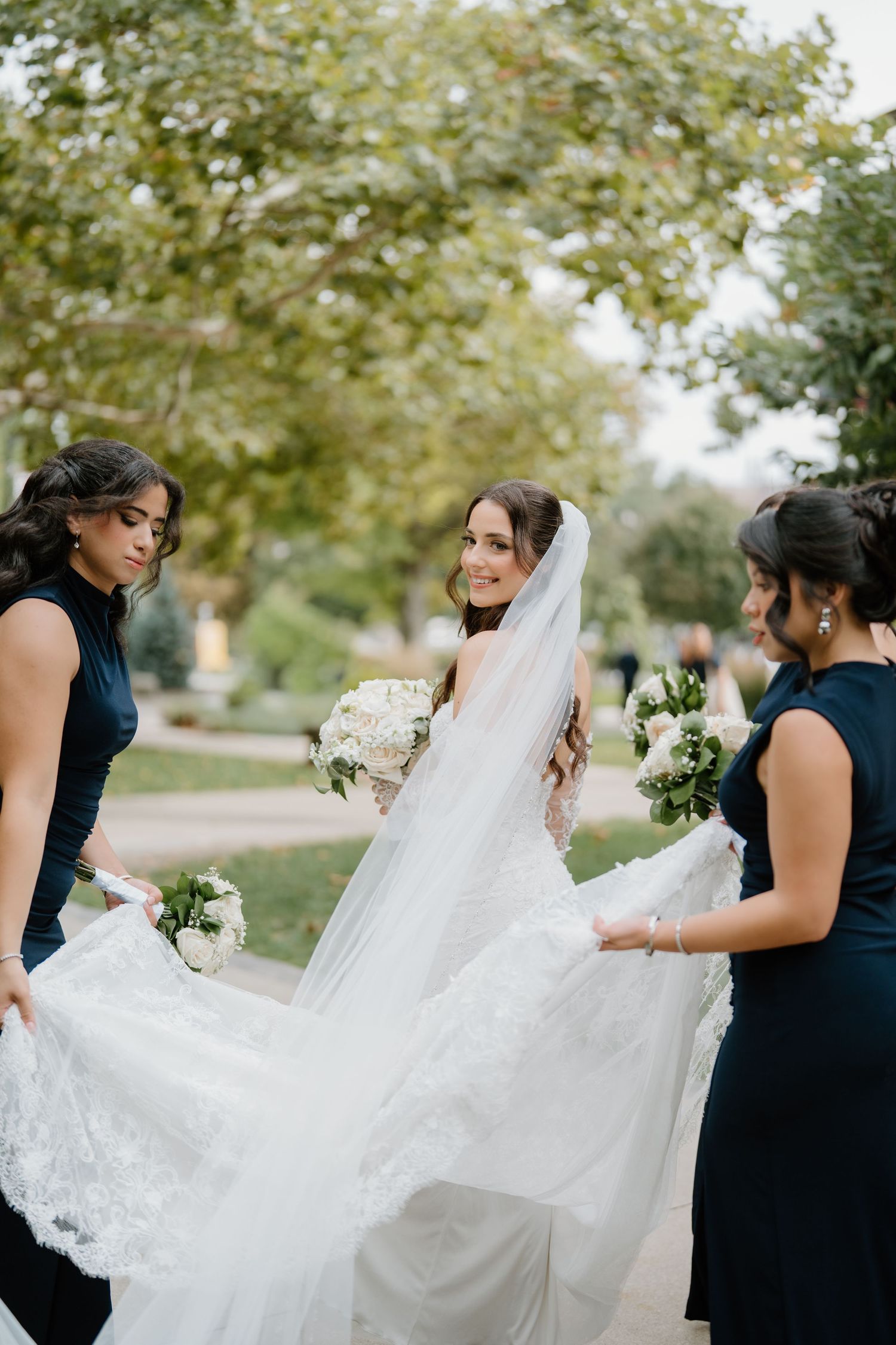 A wedding party in navy dresses helps adjust the bride's flowing white lace veil outdoors.
