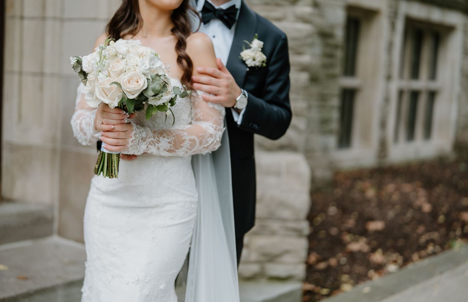 A couple shares an intimate moment in formal wedding attire against a stone building backdrop.