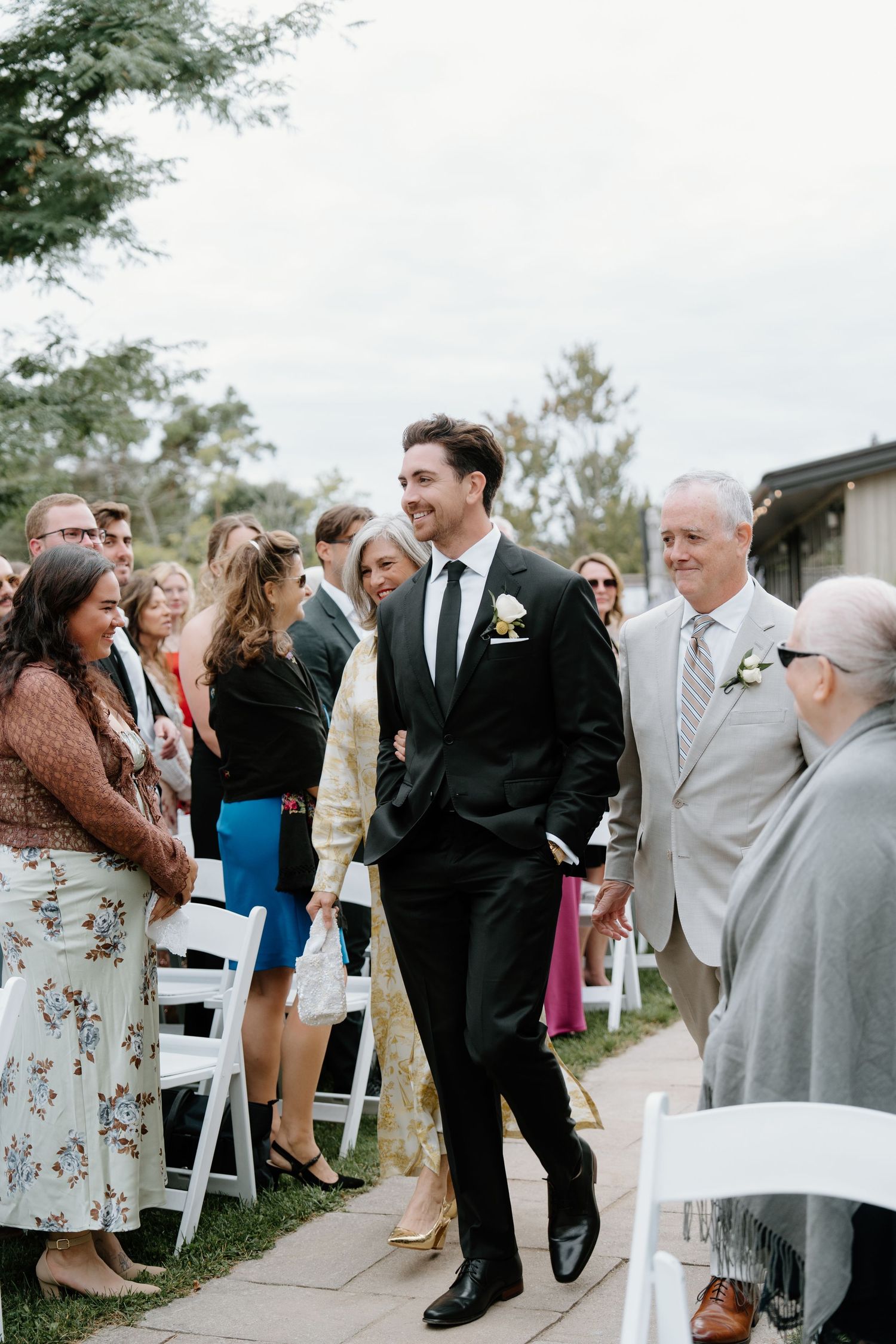 A joyful wedding ceremony moment as guests smile and gather along white chairs on a stone pathway outdoors.
