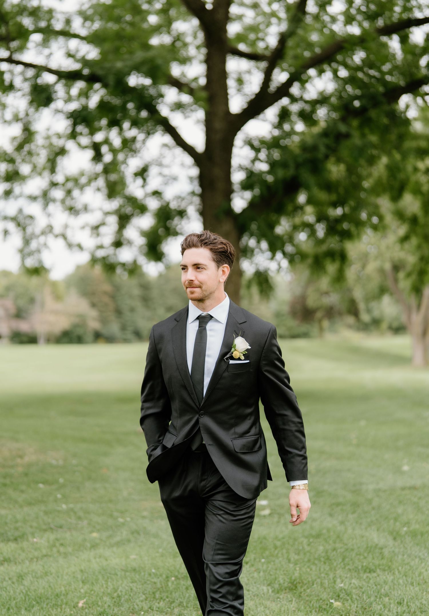 A groom in a classic black suit walks across a lush green lawn on a sunny day.