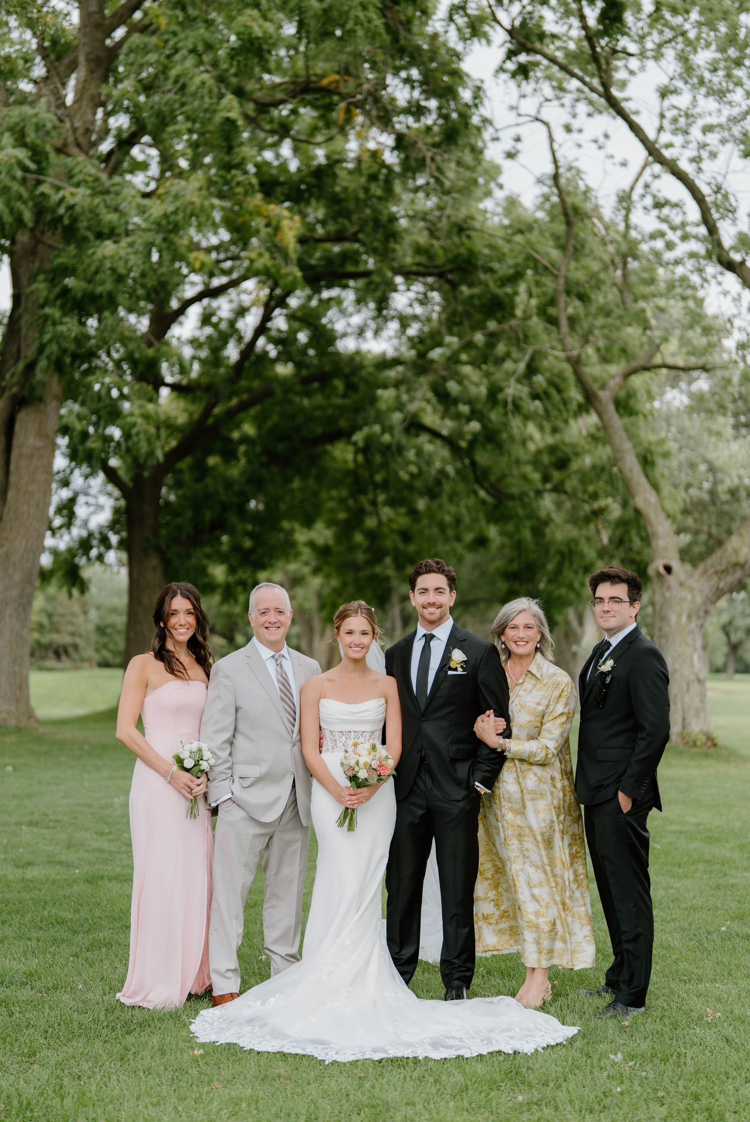 Wedding party in formal attire poses together on a grassy lawn with trees in the background.