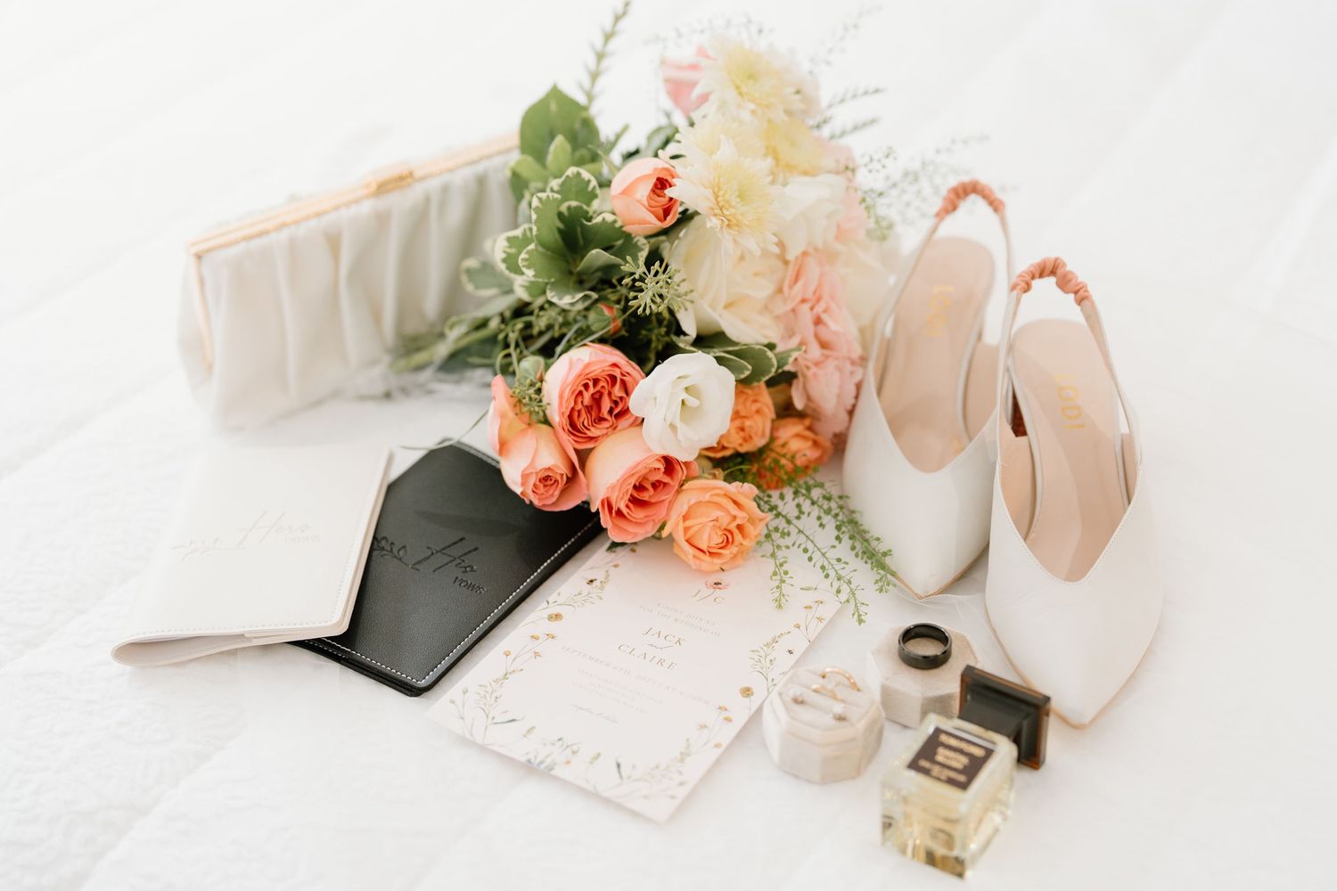 Wedding accessories including bridal shoes, floral bouquet, perfume bottle, and invitation on white marble surface.