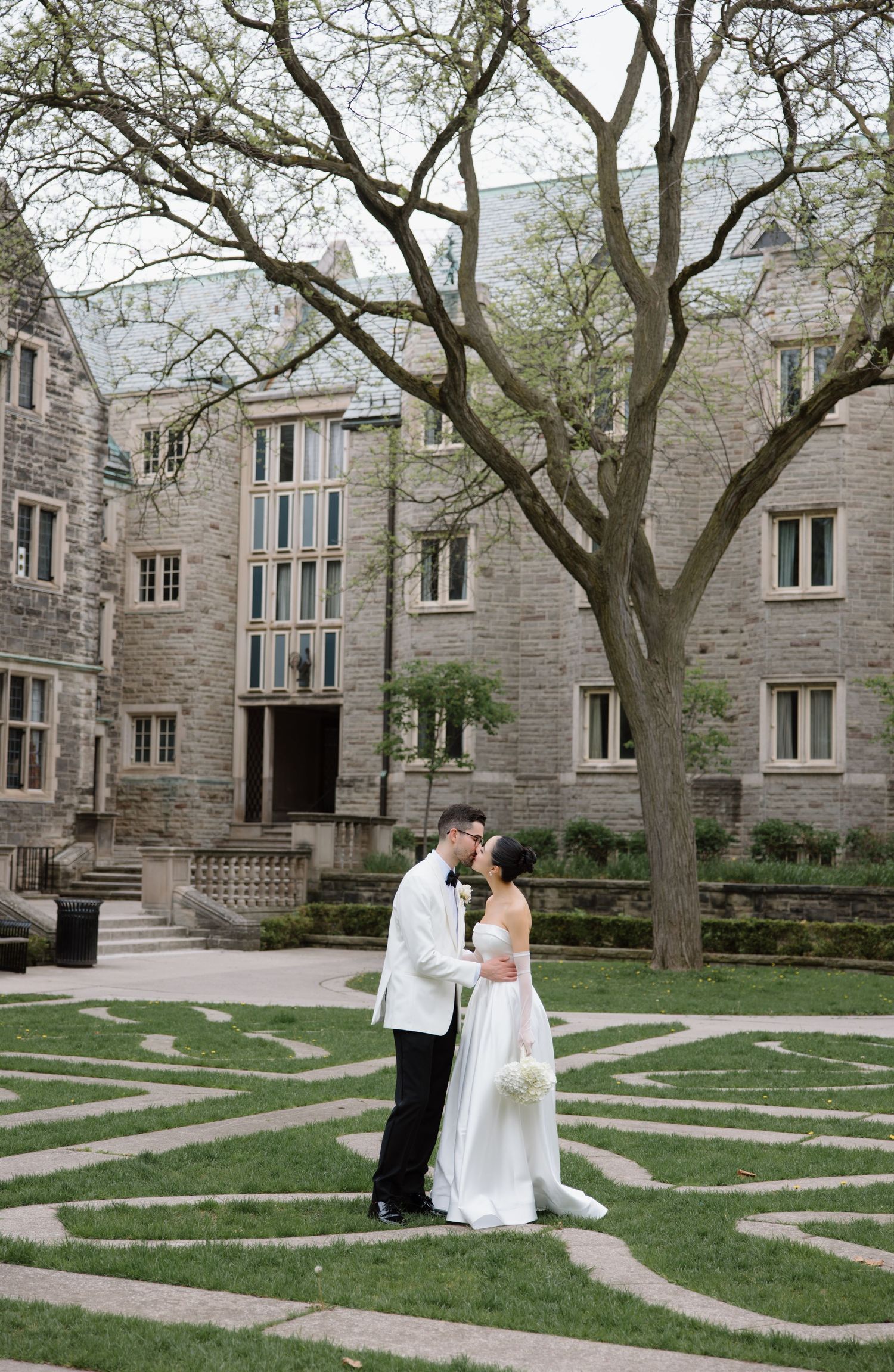 Romantic couple shares an intimate moment on a stone maze pattern lawn in front of a grand Gothic university building.
