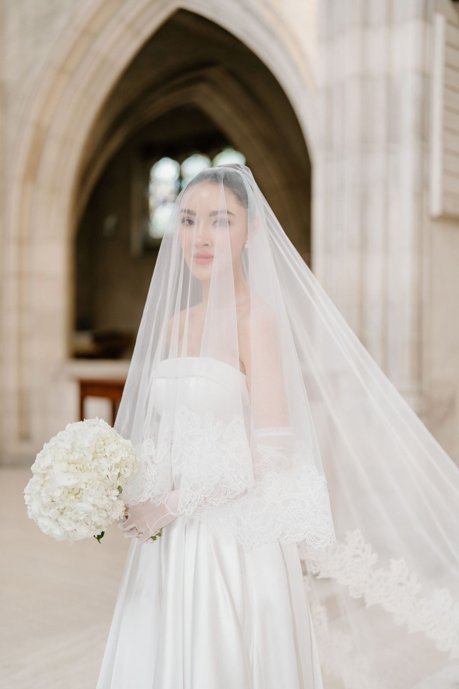Elegant bride in strapless white dress and flowing veil poses in stone archway with white hydrangea bouquet.