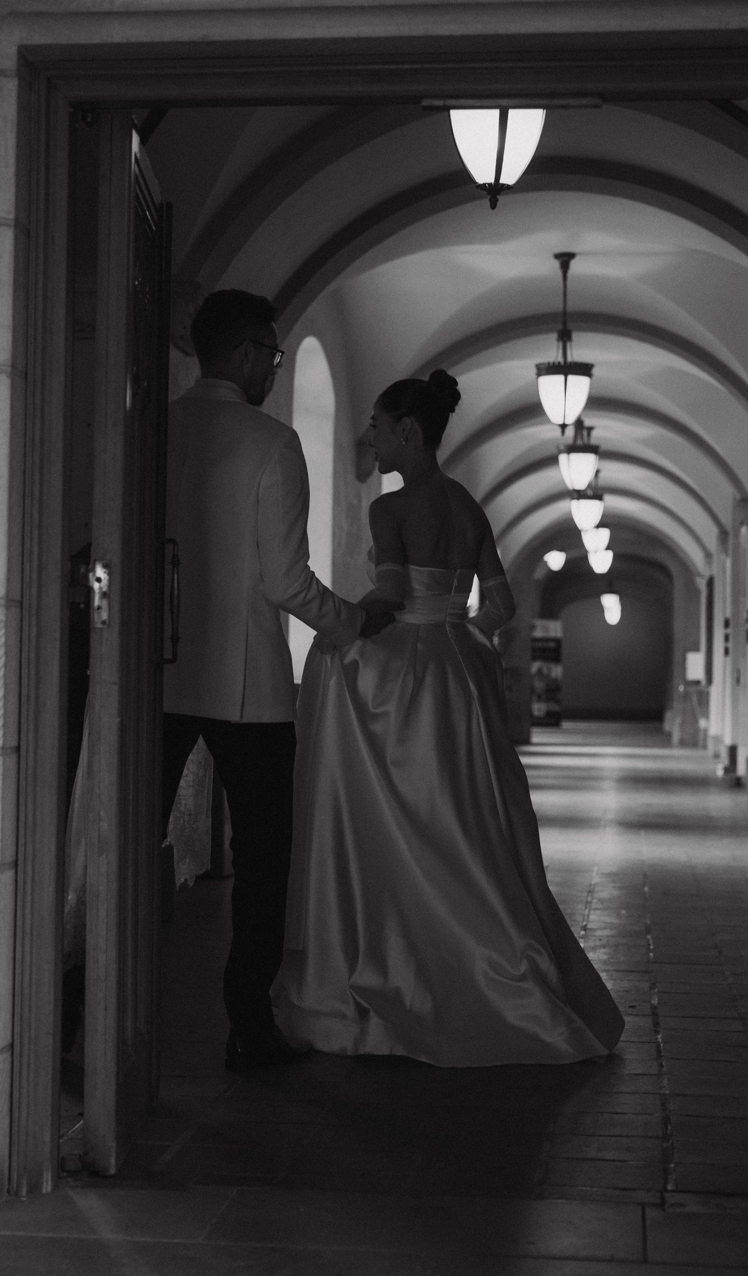 A couple in formal attire stand in an elegant arched hallway illuminated by vintage pendant lights in black and white.