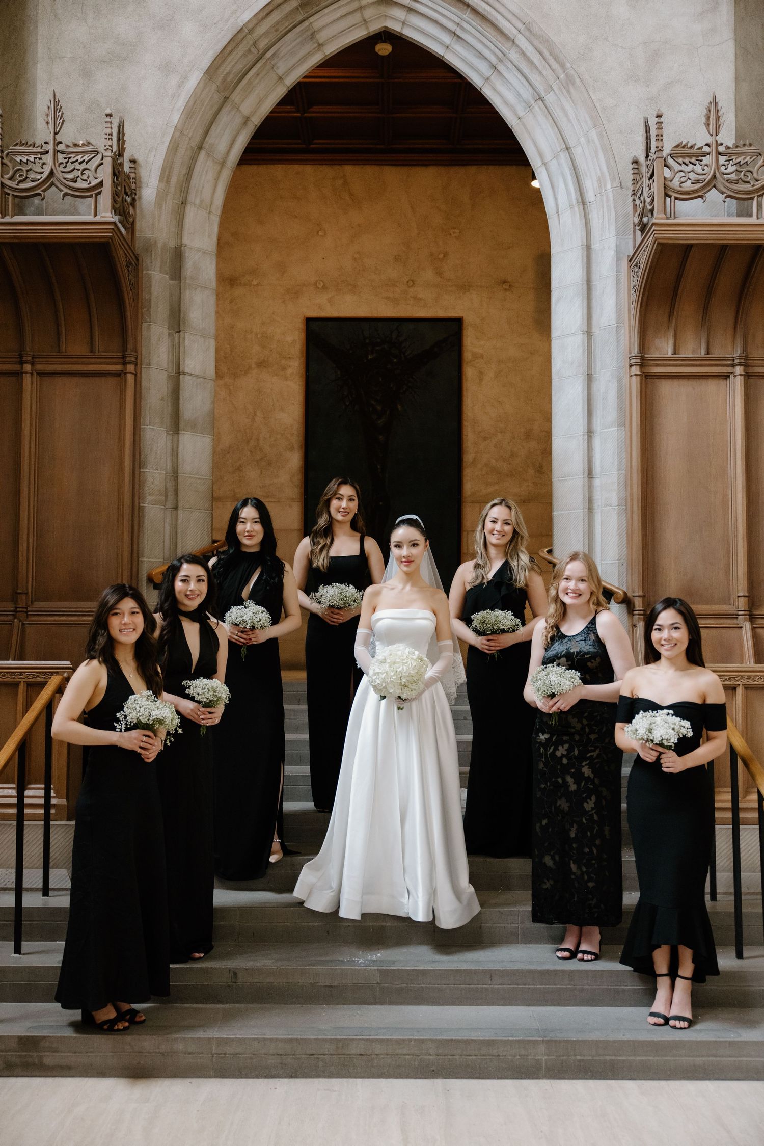 A wedding party in black dresses and a bride in white pose together under elegant stone archways.