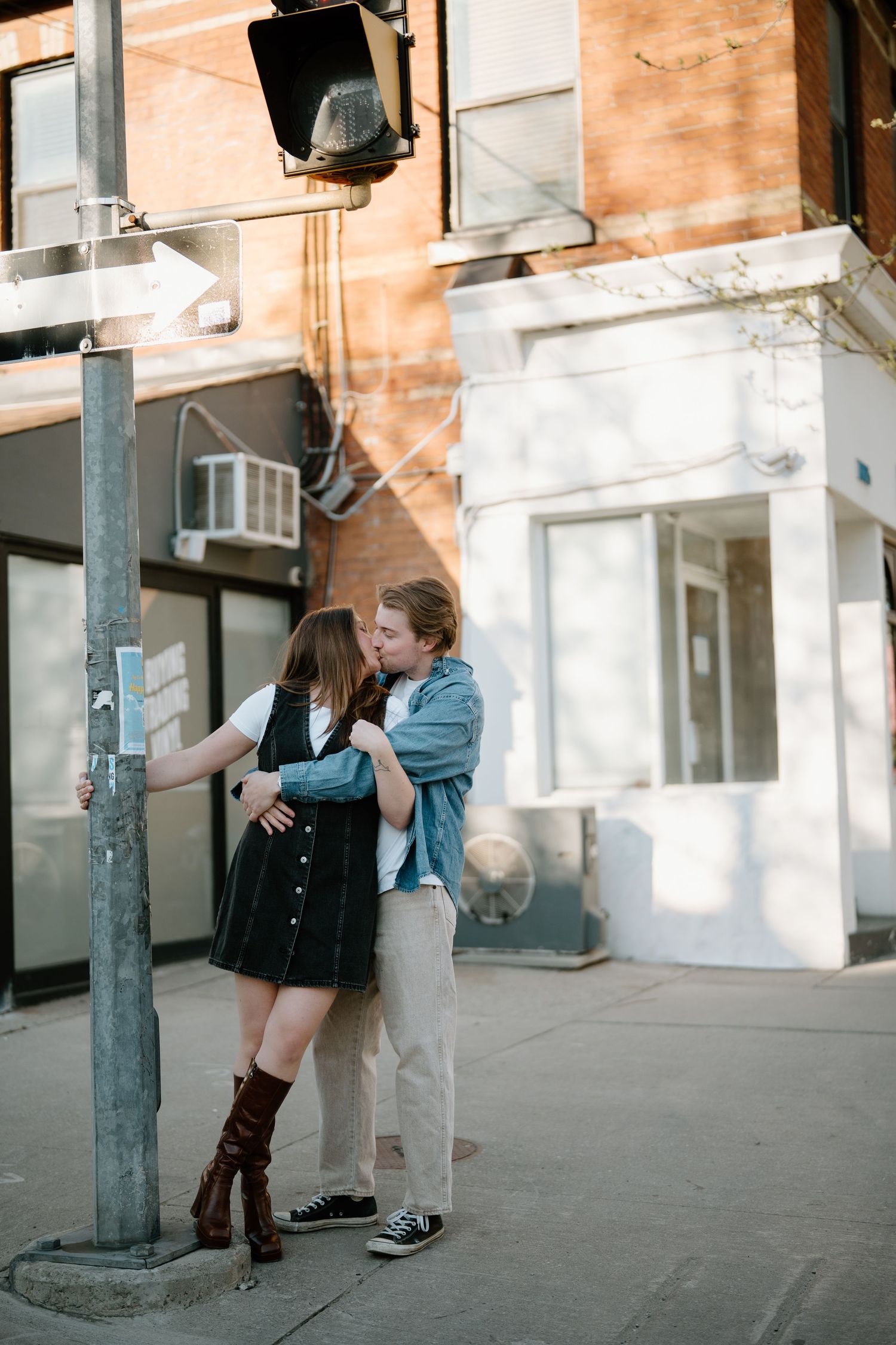 A couple embraces on an urban street corner under a hanging lamp on a sunny day.