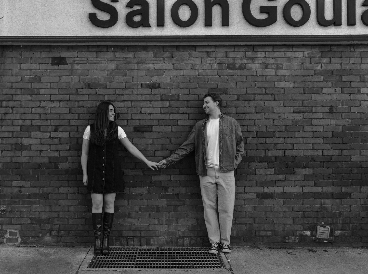 A couple holds hands while standing against a brick wall with a salon sign above in black and white.