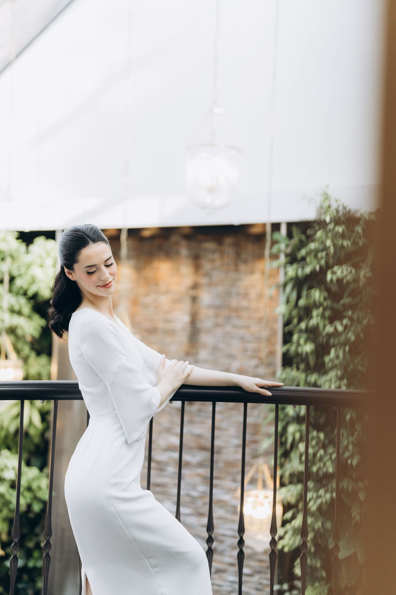 Elegant figure in flowing white dress poses gracefully on a balcony with brick wall and greenery in background.