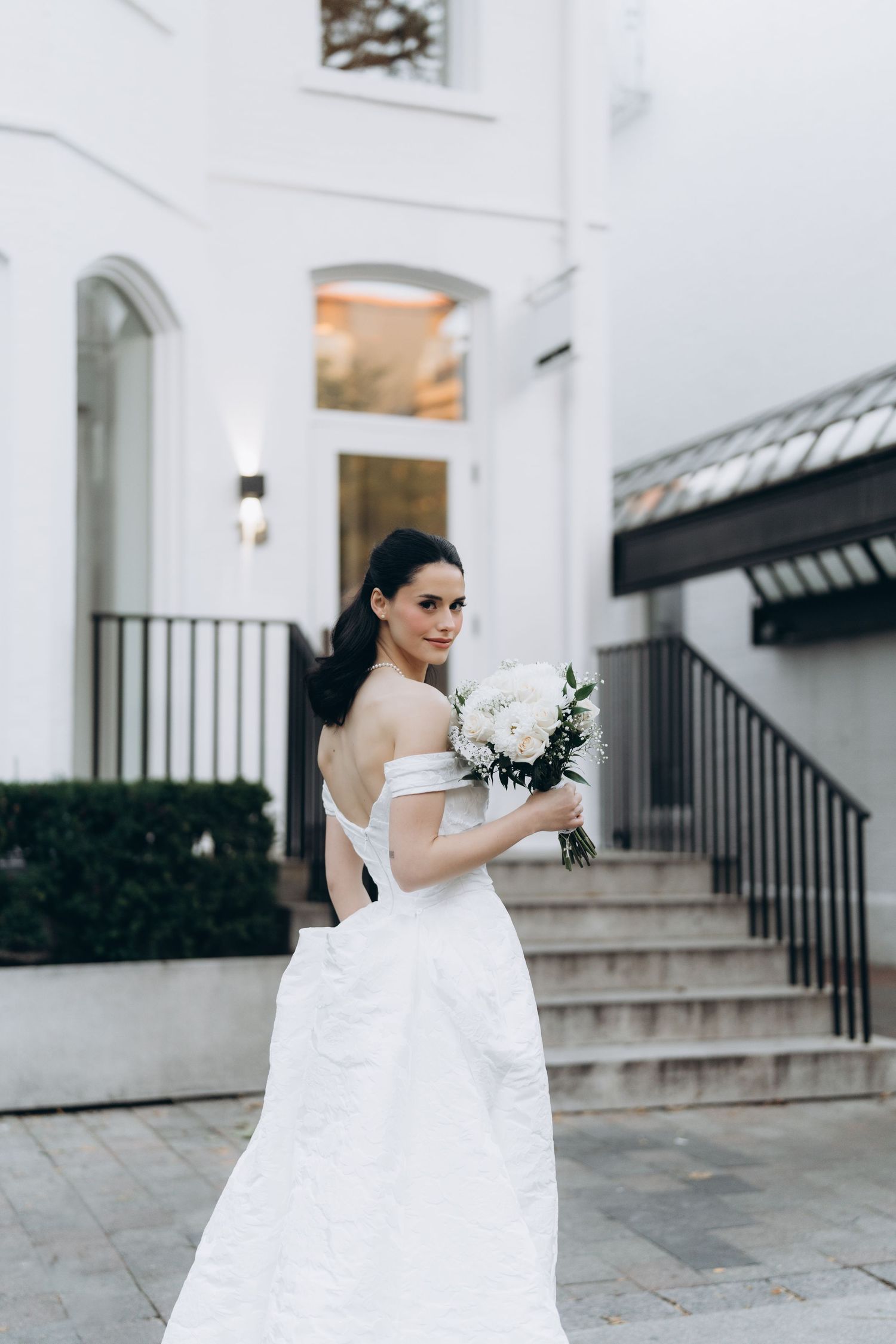 A bride in a white wedding dress stands elegantly against a white building exterior with black railings.