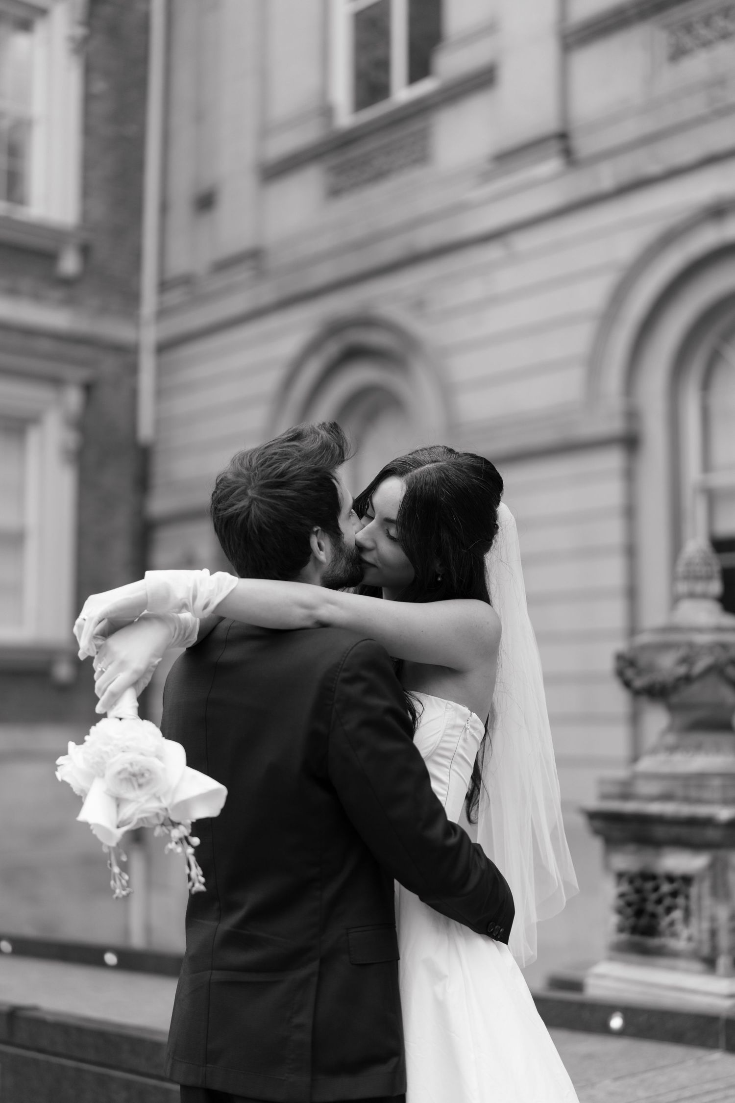 A bride and groom share a romantic embrace outside a historic building with elegant architecture in black and white.