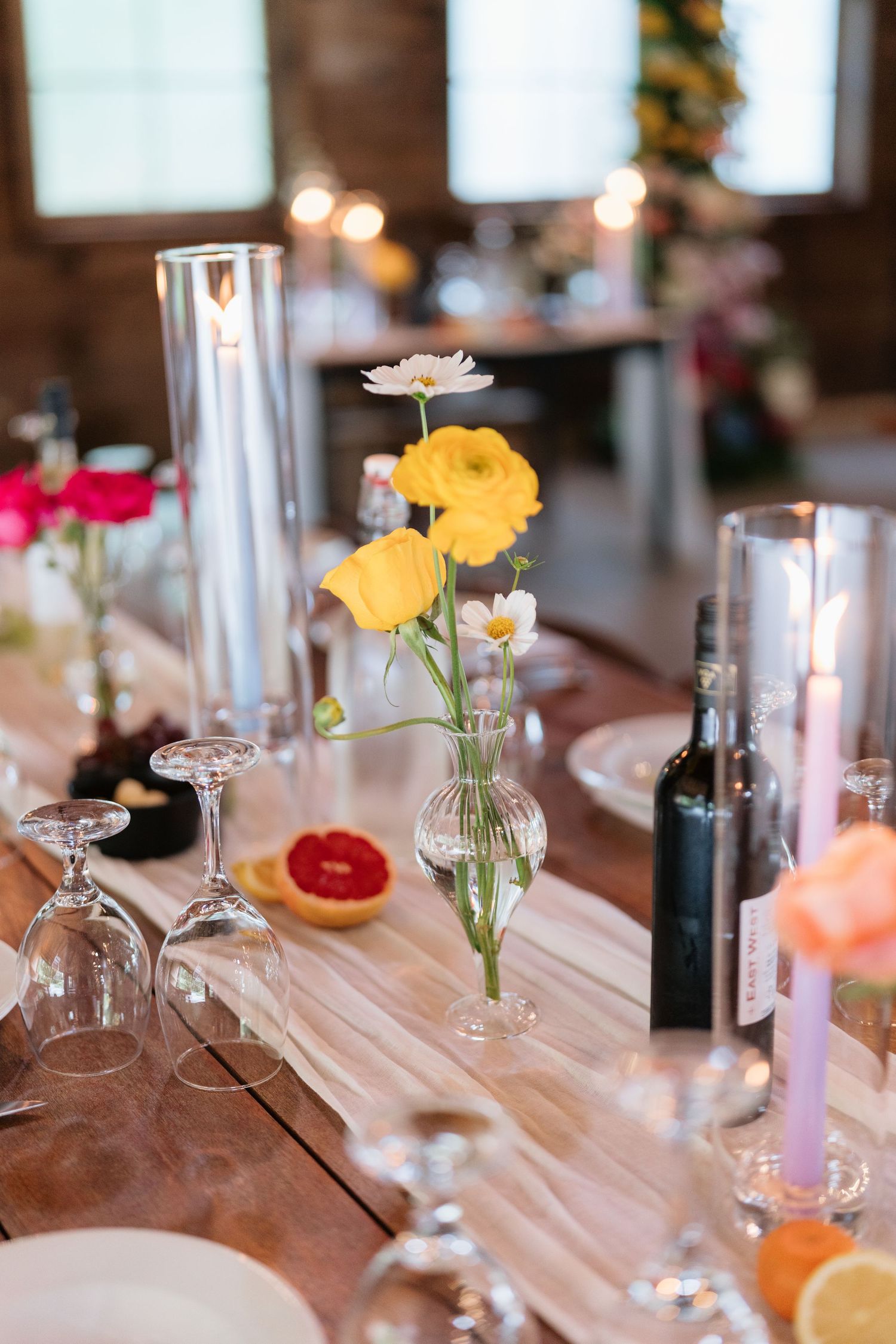 Elegant table setting with yellow ranunculus flowers in glass vase, wine glasses and citrus accents on wooden surface.