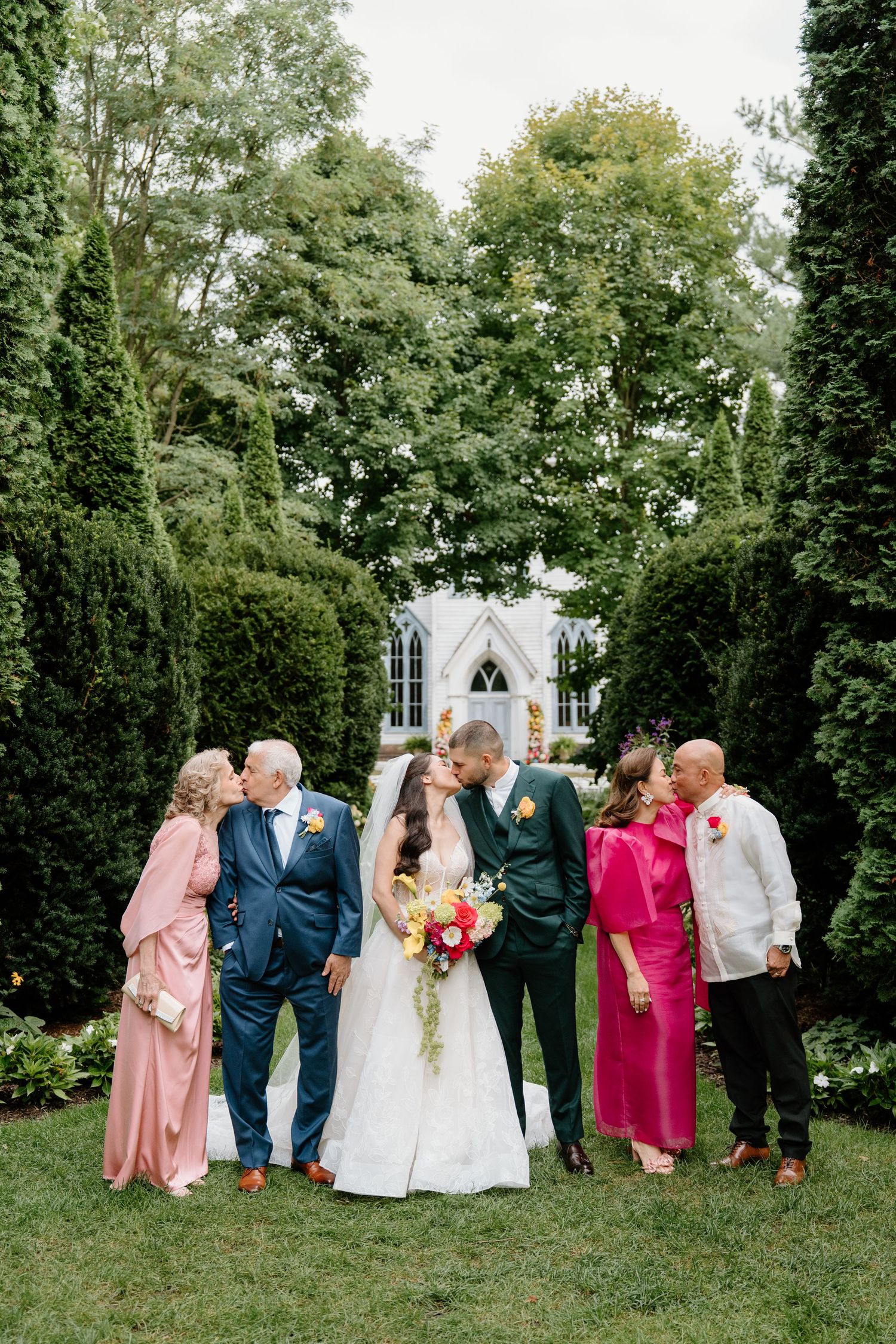 Wedding party poses for creative leaning photo series in garden setting with lush green trees in background.