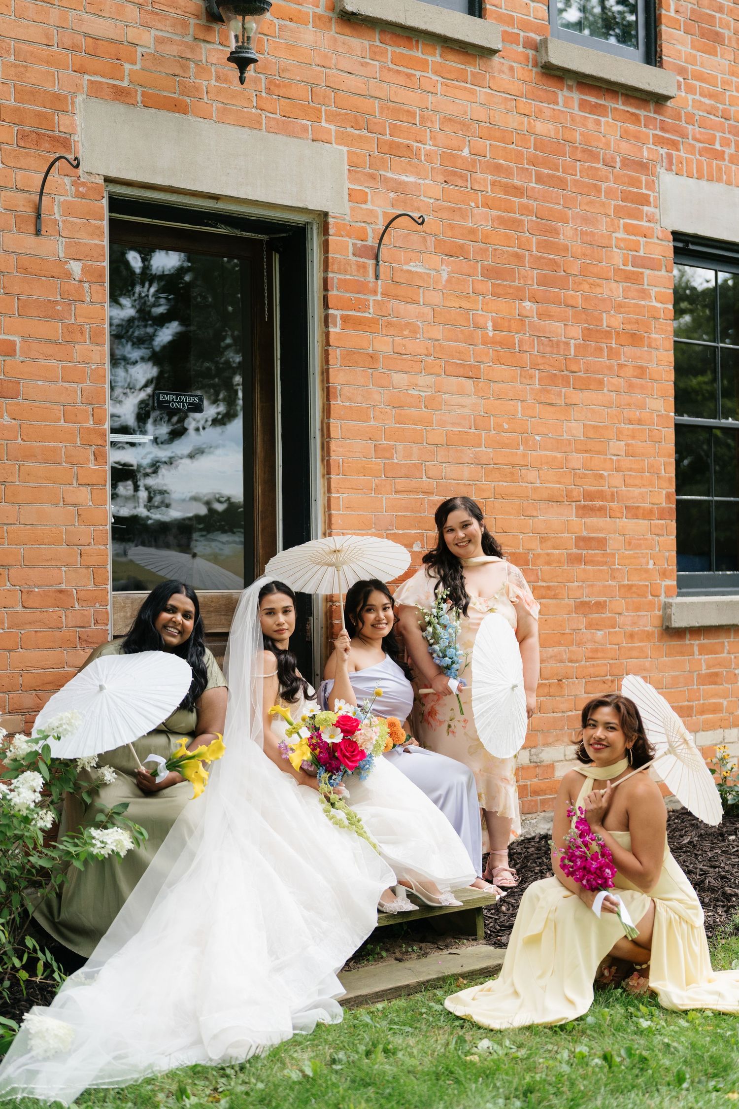 Wedding party in yellow dresses and white gowns pose creatively along a brick building exterior.
