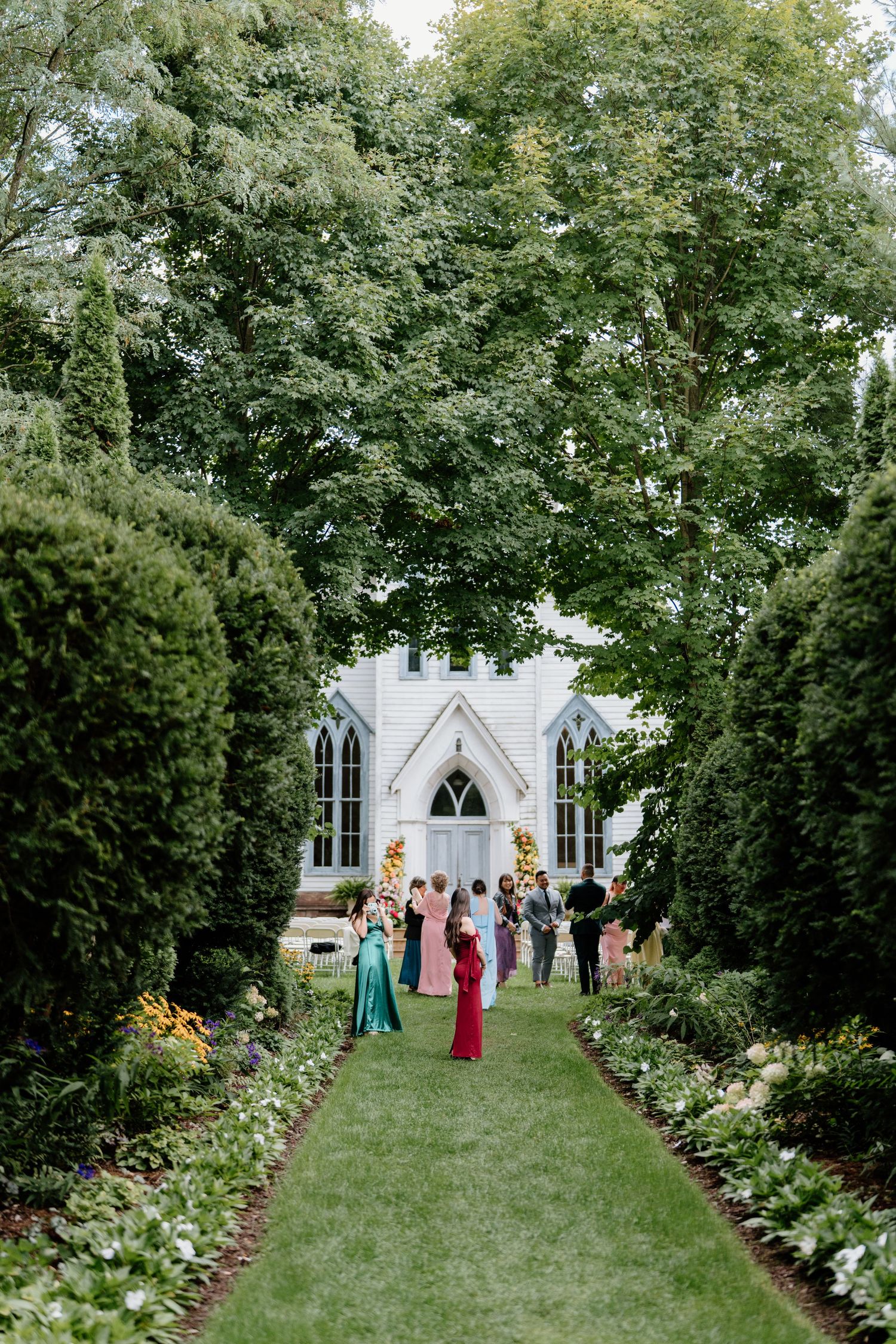 A wedding ceremony taking place in a lush garden setting with a white gothic chapel and guests gathered on a grass pathway.