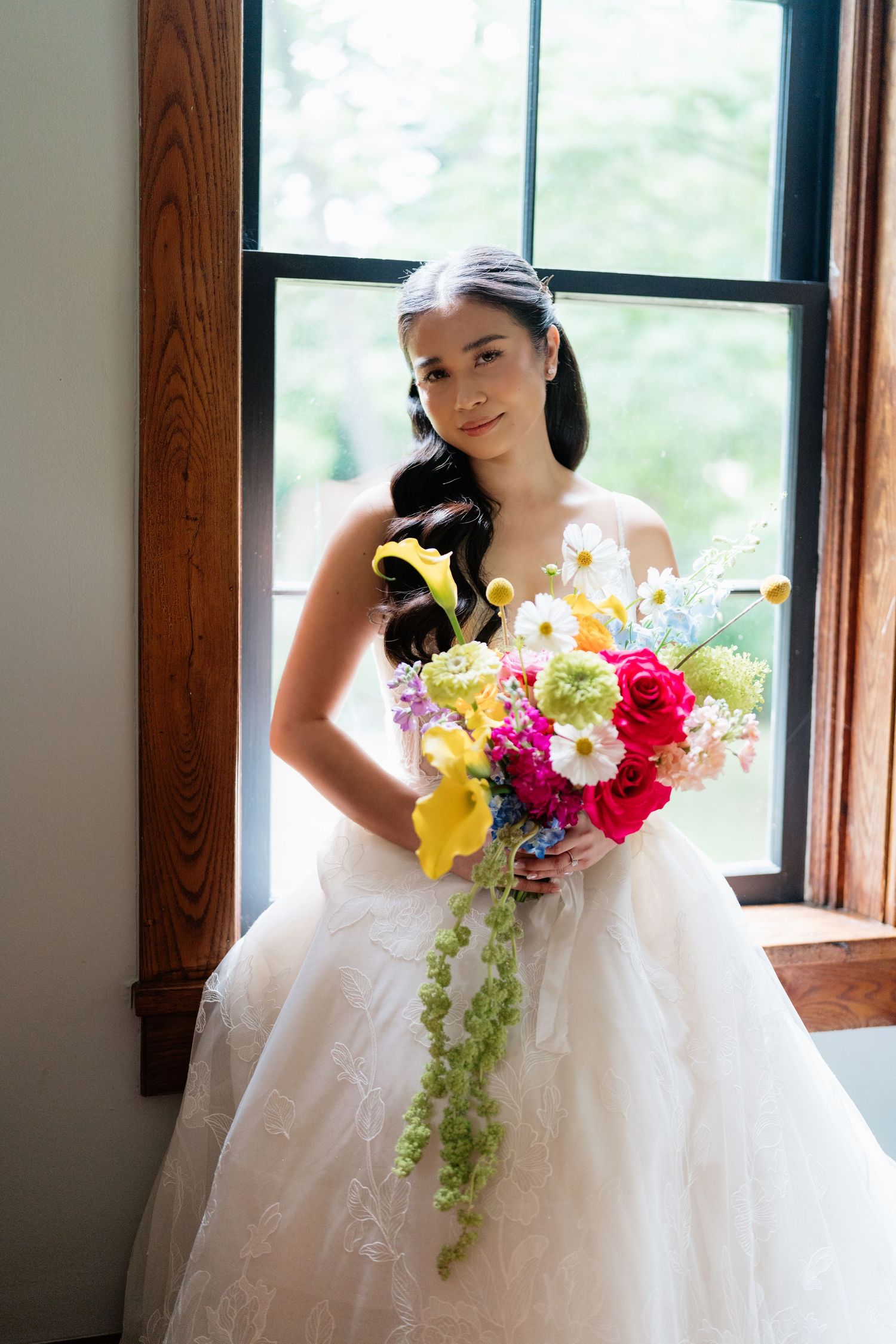 A bride in a white wedding gown holds a vibrant bouquet of colorful flowers near a sunlit window.