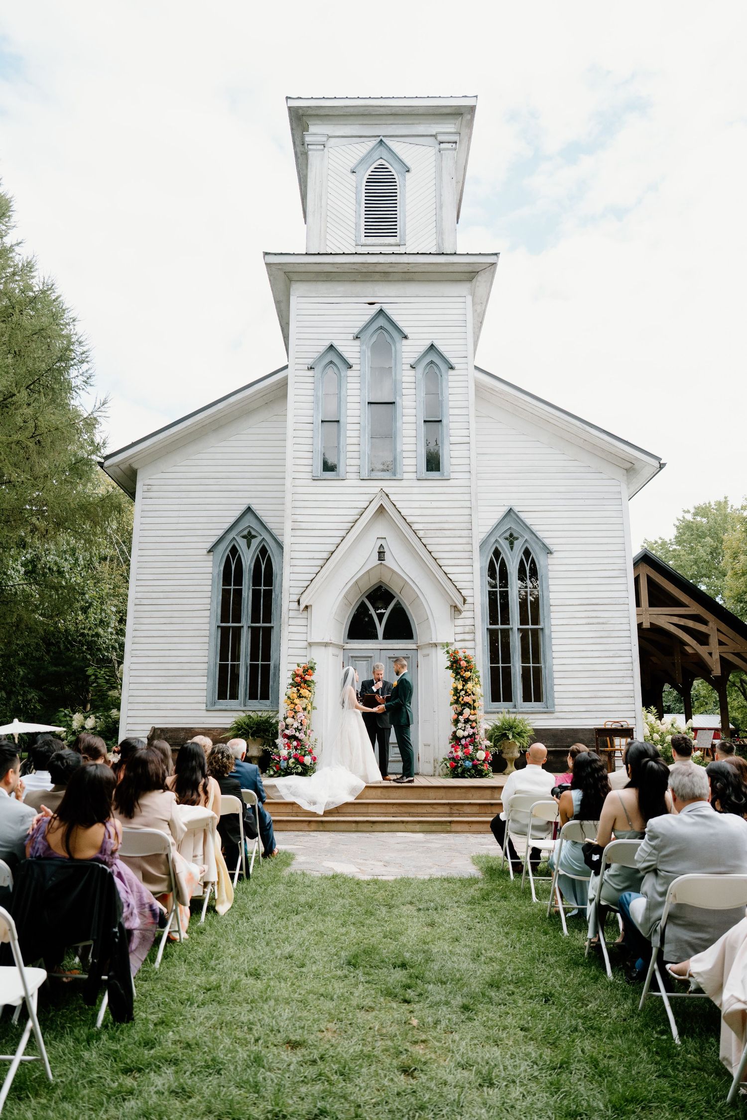 An outdoor wedding ceremony at a charming white historic church with guests seated on folding chairs on the lawn.