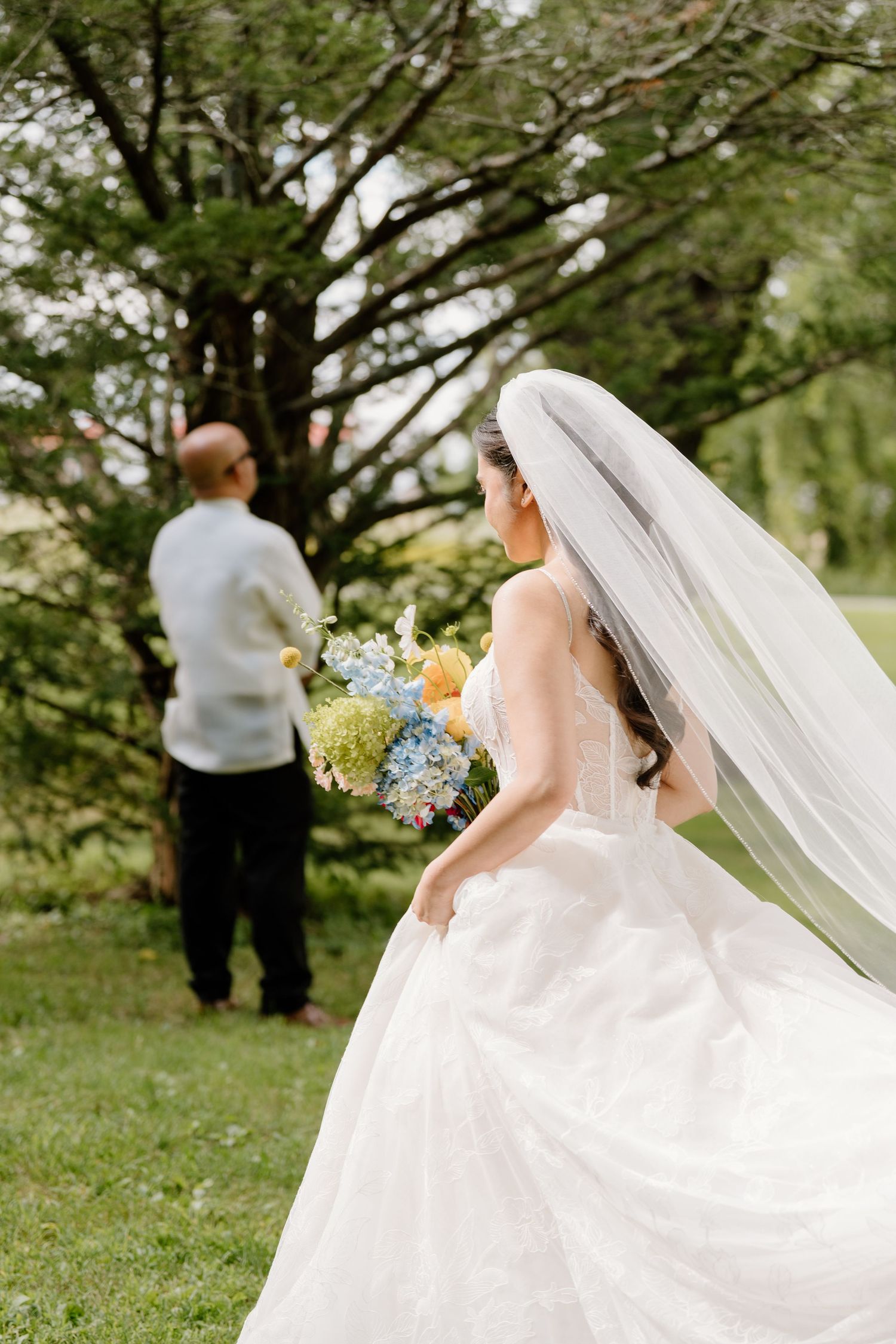 A flowing white wedding dress and veil flutter in the breeze as figures walk through a lush green garden setting.