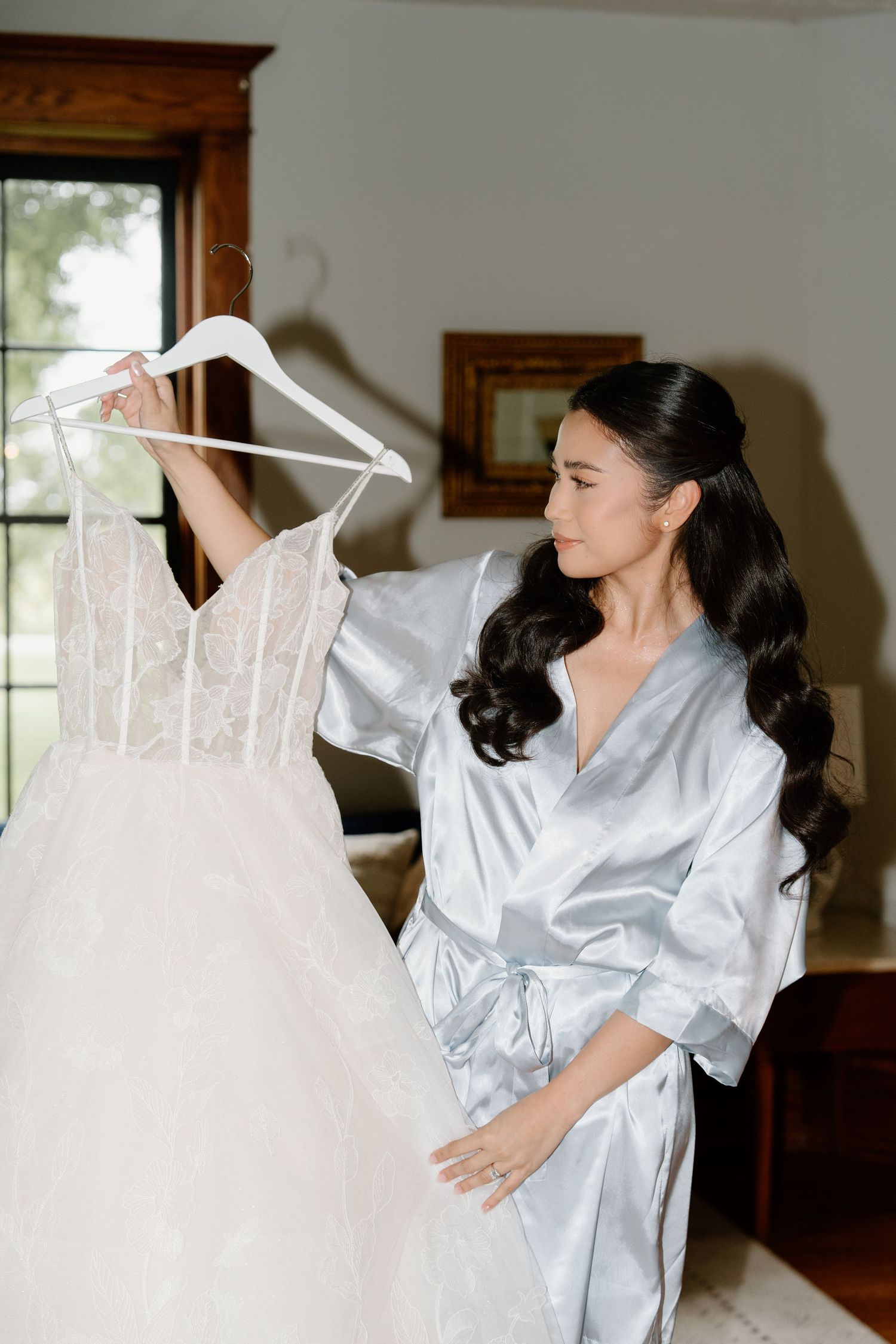 A white wedding dress on a hanger being admired by someone wearing a light blue silk robe in a sunlit room.