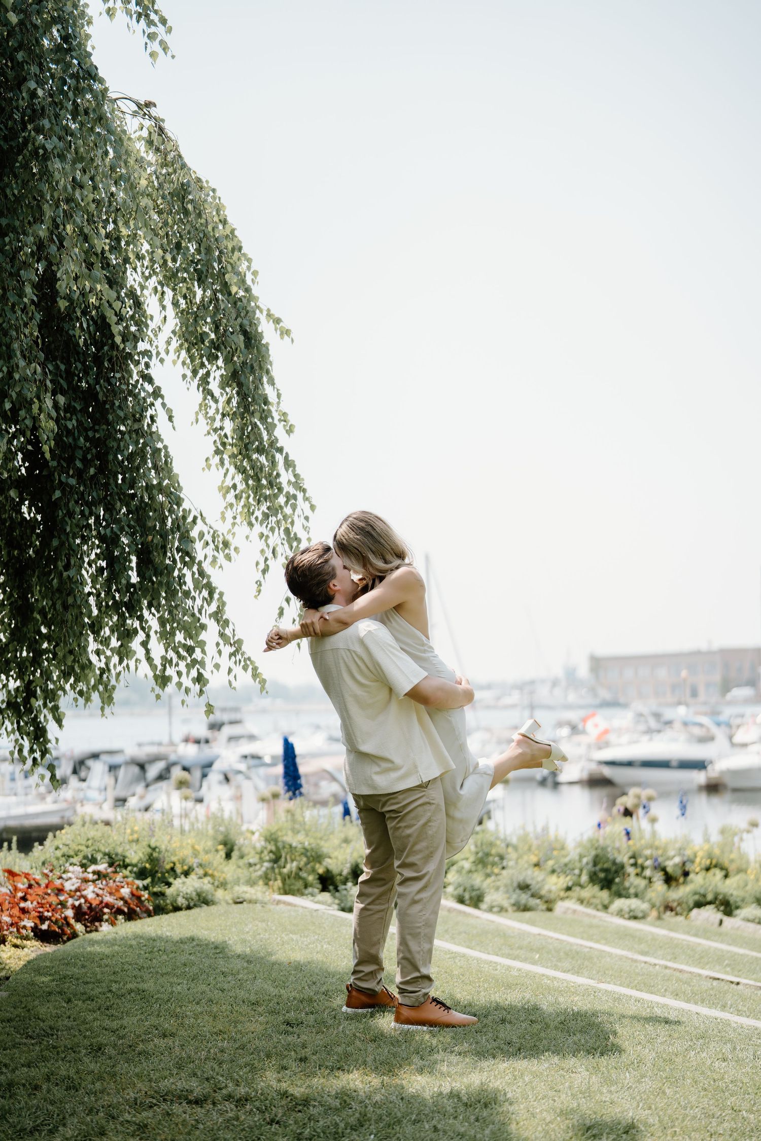 A romantic couple in light-colored attire shares an intimate embrace on a grassy lawn near a marina.