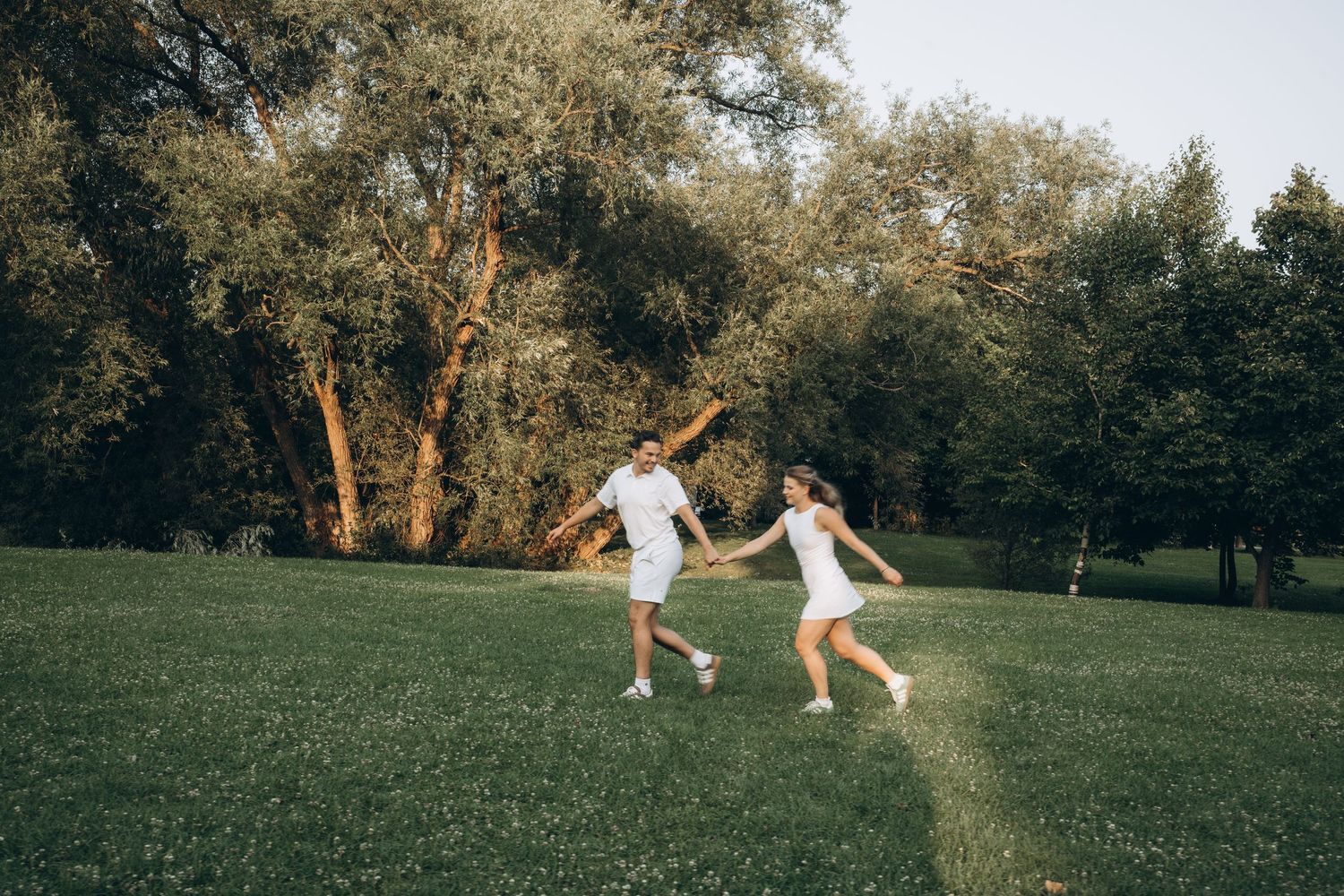 Two people in white dresses dance joyfully on a grassy field surrounded by tall trees on a summer evening.