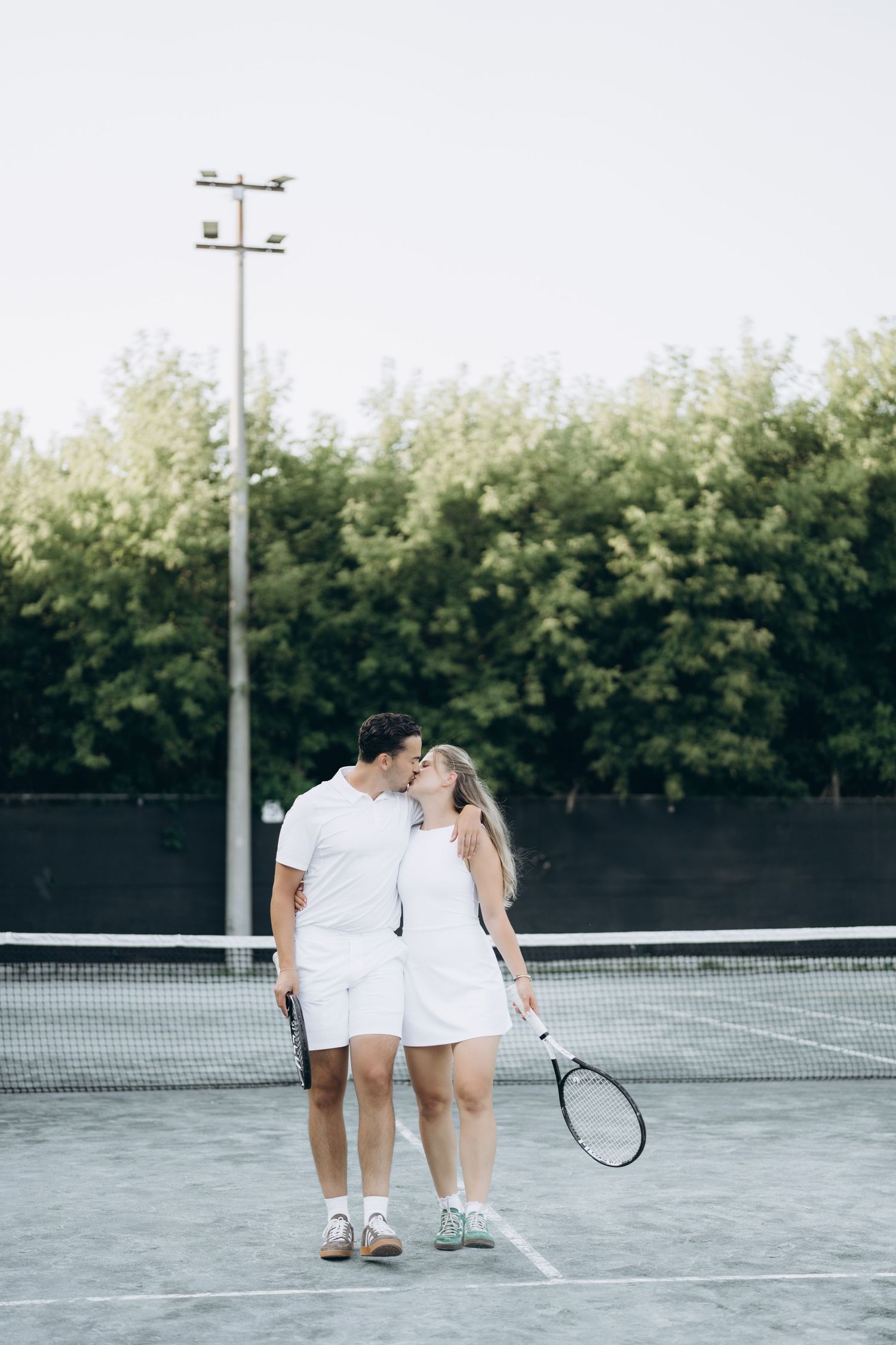 A couple in white tennis outfits share a romantic moment on an outdoor tennis court.