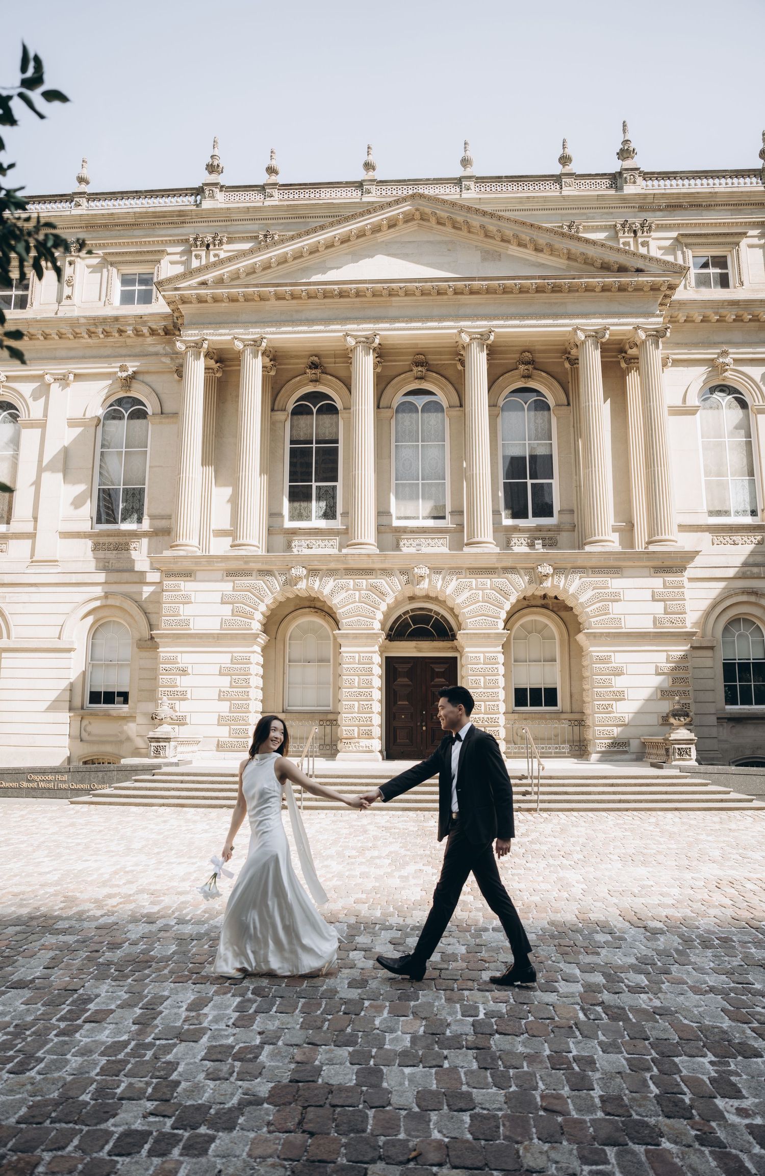 A couple in wedding attire walks on cobblestones in front of an elegant neoclassical building with grand columns.