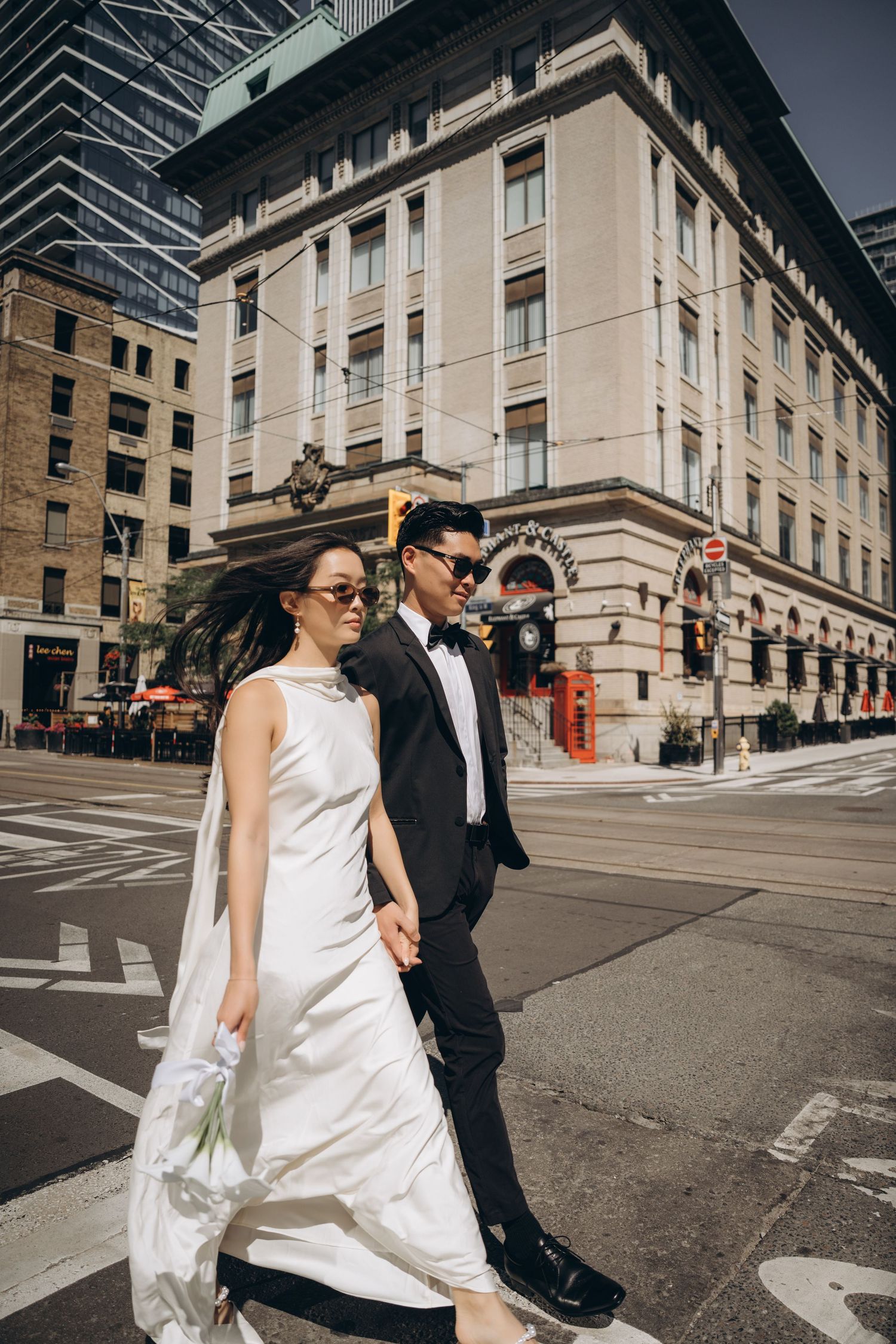 A couple in wedding attire walks across an urban intersection with sunlit city buildings in the backdrop.