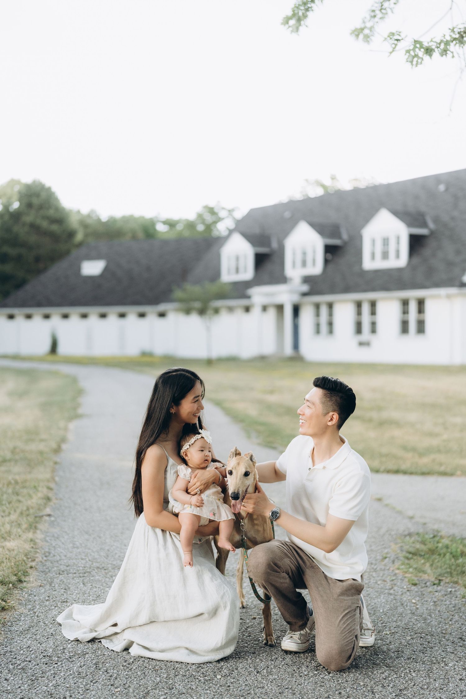 A family sits on a path with their whippet dog in front of a white house with dormers.
