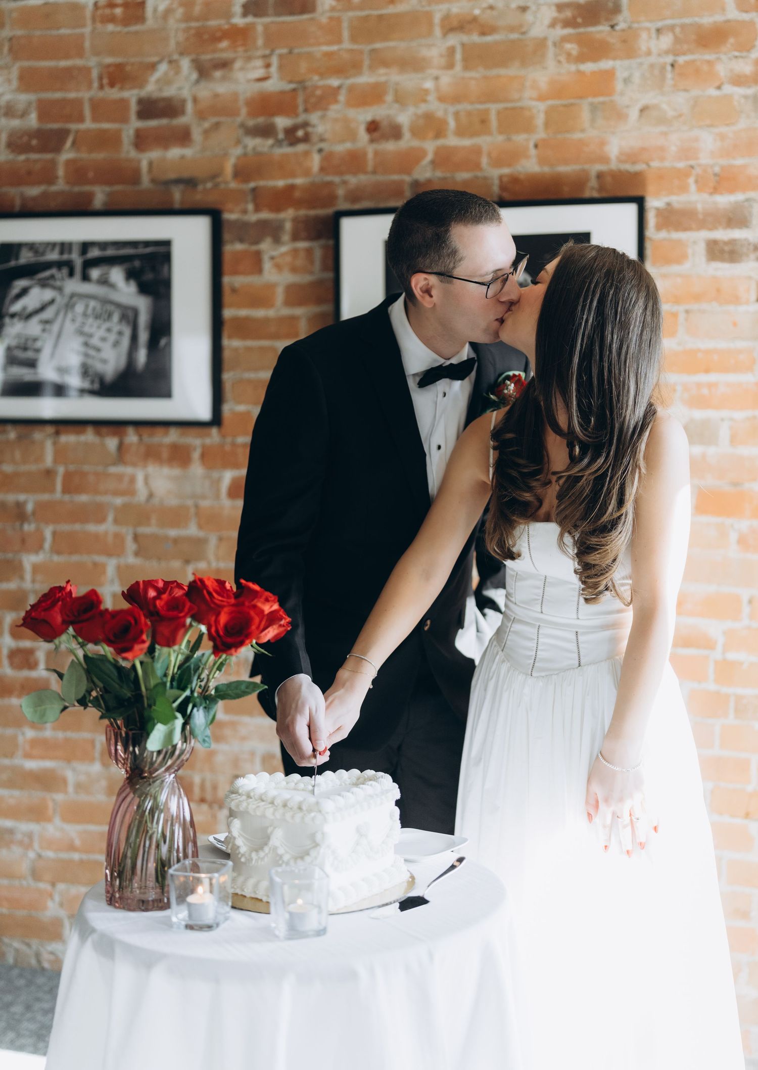 Romantic wedding cake cutting moment in rustic brick venue with red roses and white decor.