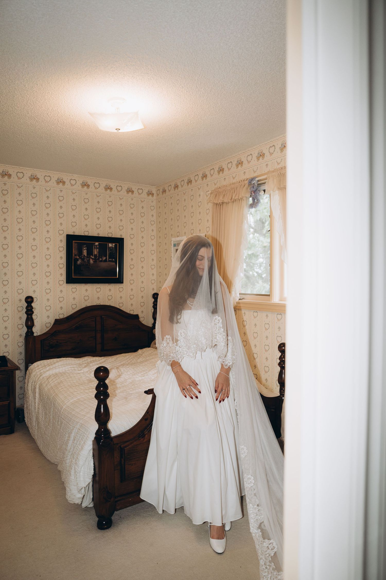 A white wedding dress displayed in a bedroom with a dark wooden four-poster bed and floral wallpaper.