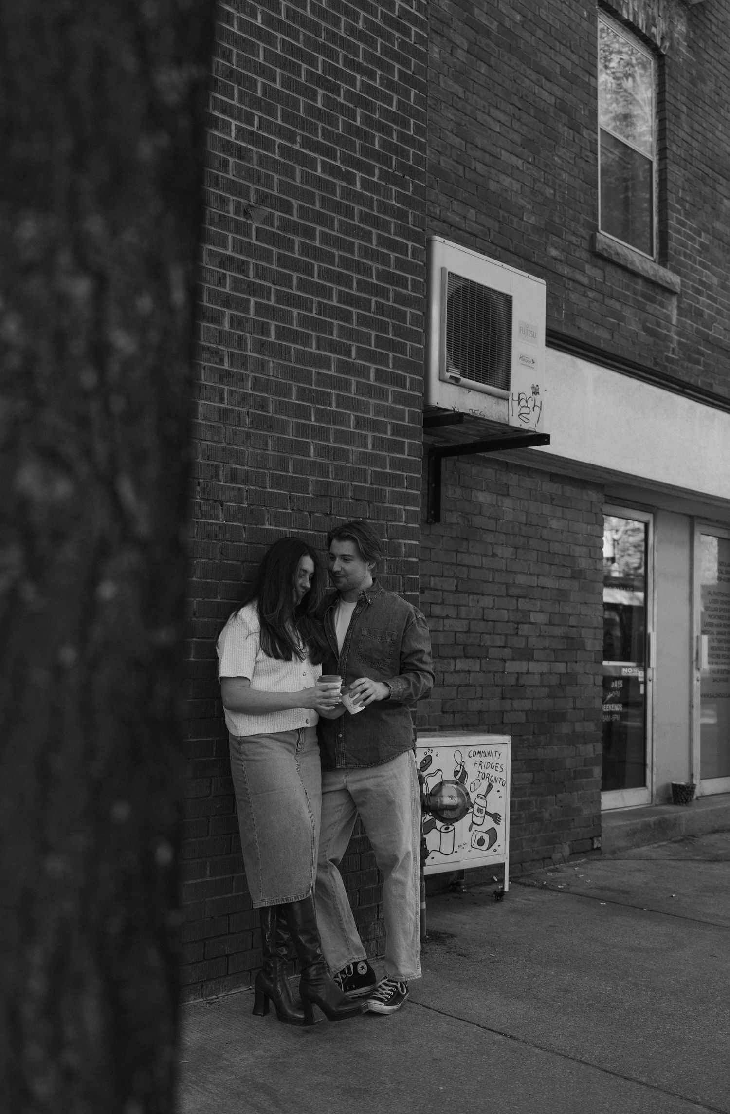 A couple stands intimately against a brick wall outside a storefront in a black and white photograph.