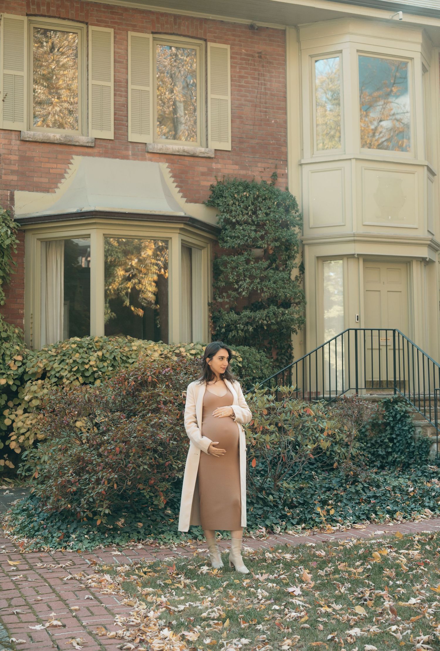 An elegant fall scene features a brick house with bay windows and manicured shrubs along a brick walkway lined with leaves.