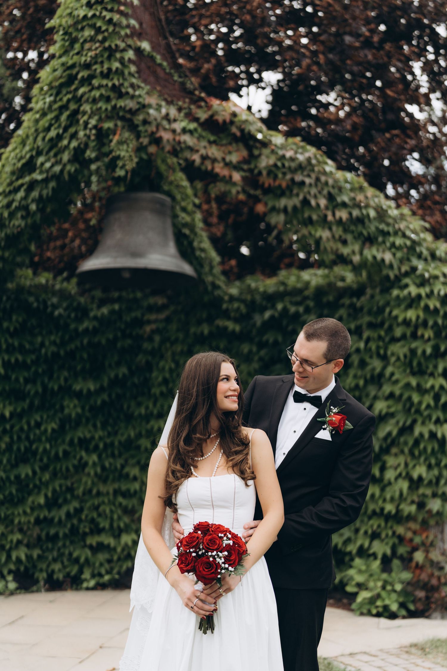 A couple poses together during their wedding photoshoot in front of green foliage wearing formal attire.