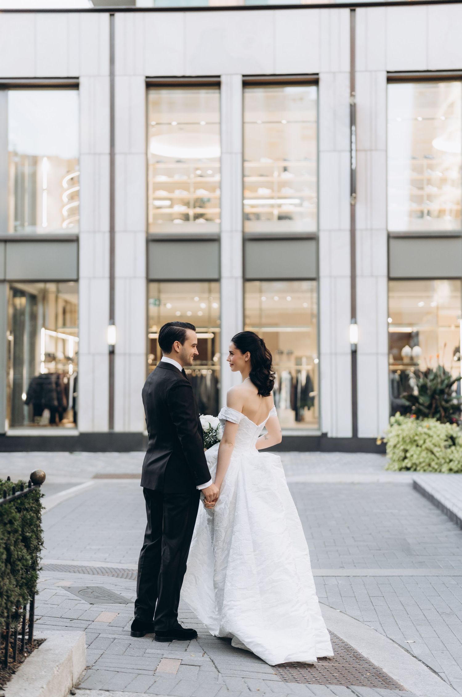 A bride and groom share a tender moment on a modern patio with large windows and architectural columns.