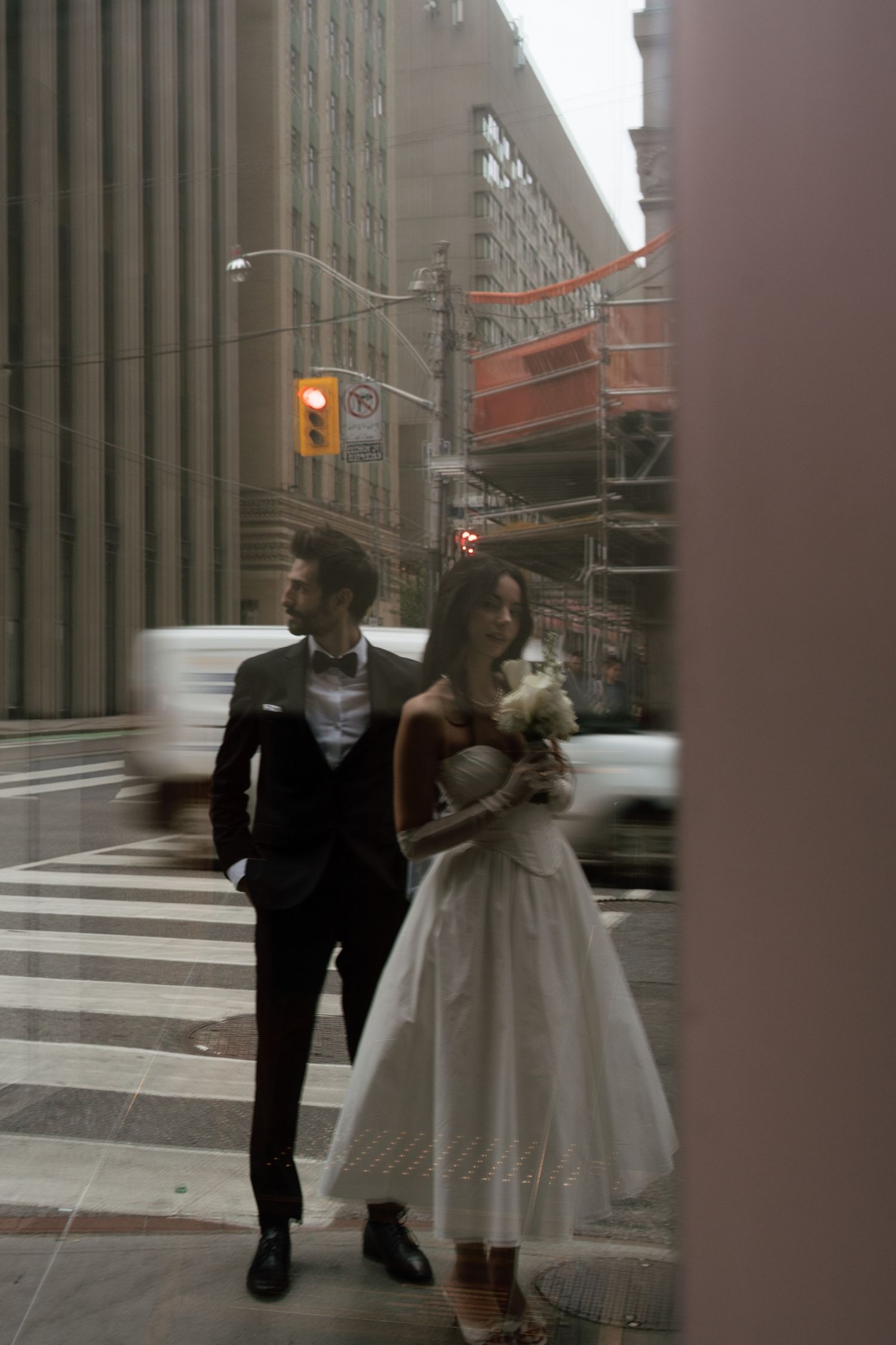 A bride and groom walk together through a city street with tall buildings in the background on a cloudy day.