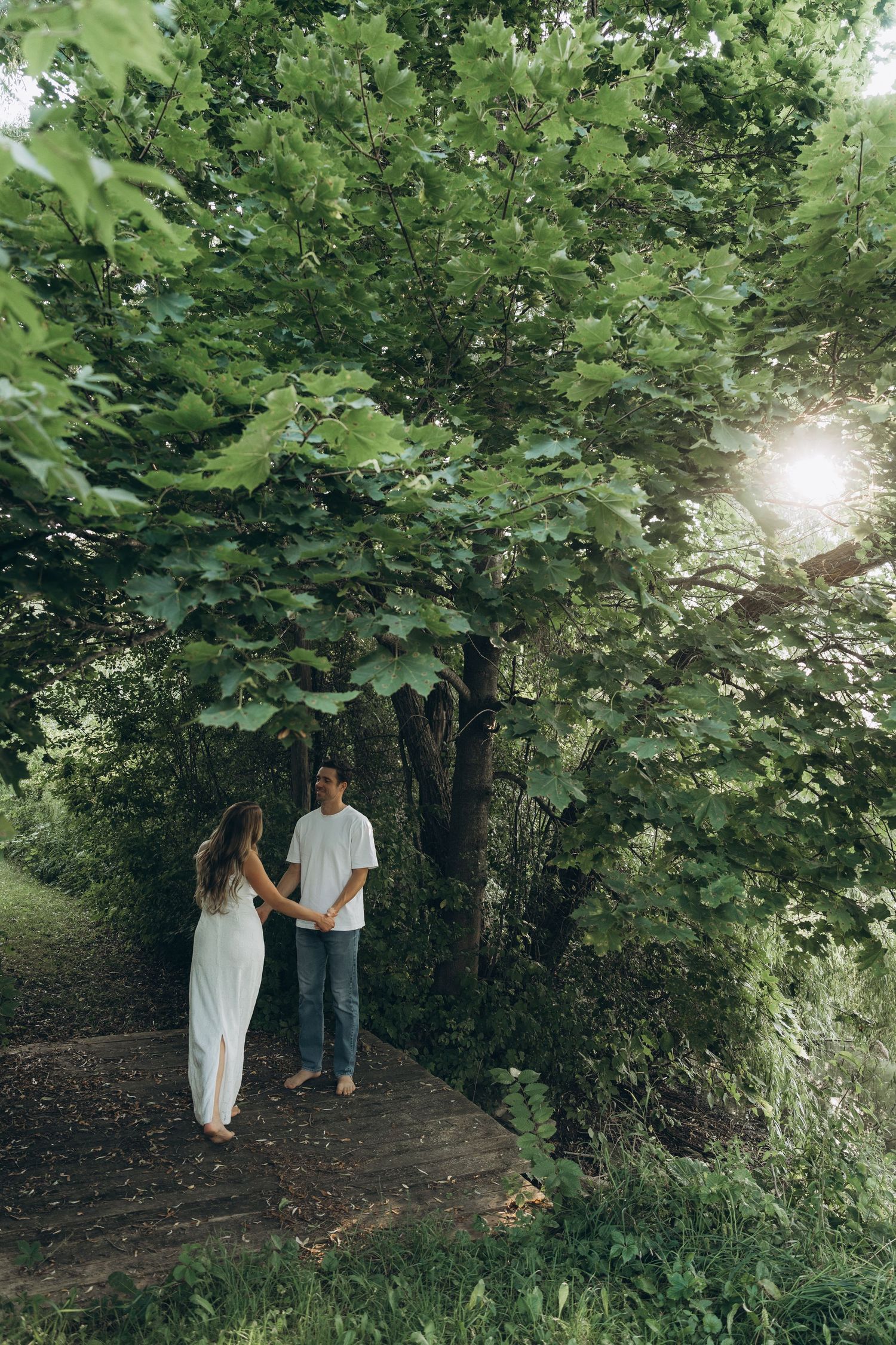 A couple stands together on a wooded path surrounded by lush green foliage as sunlight filters through the trees.