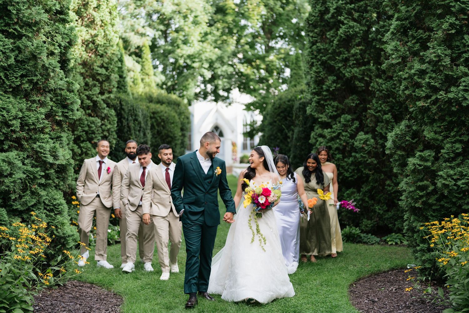 Wedding party in formal attire poses together in a lush garden setting during summer celebration.