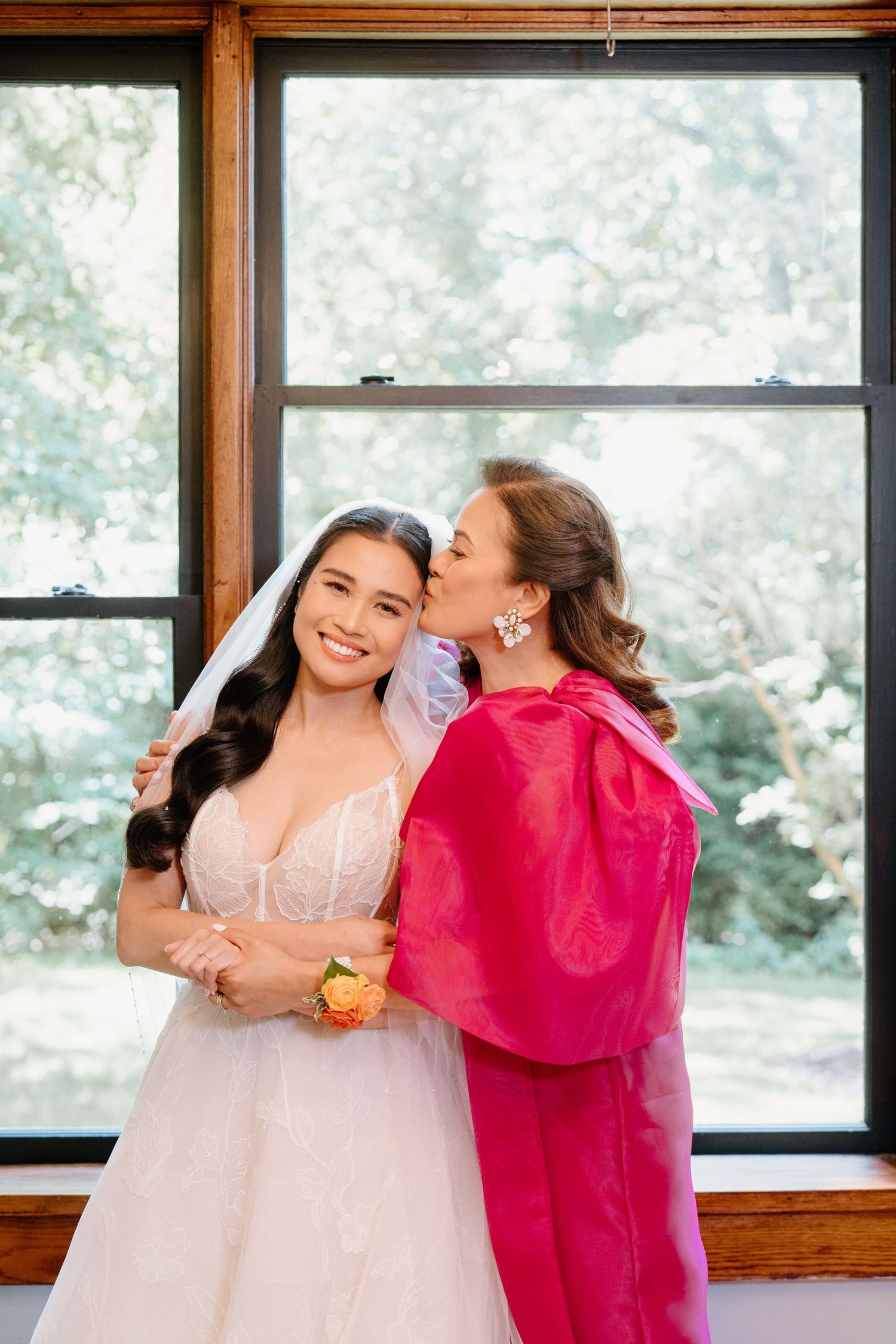 Two people embrace and laugh together, one in a white dress and another in a vibrant pink outfit near windows.