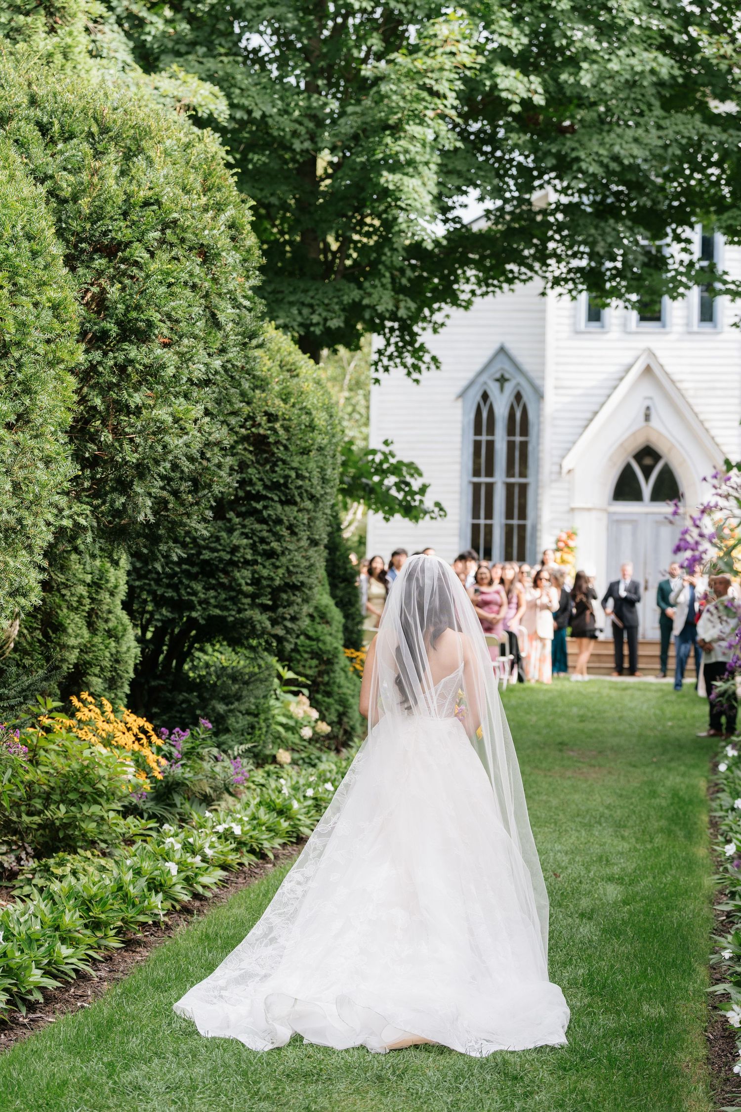 A bride in a flowing white gown and veil walks along a garden path toward a white church with guests gathered outside.
